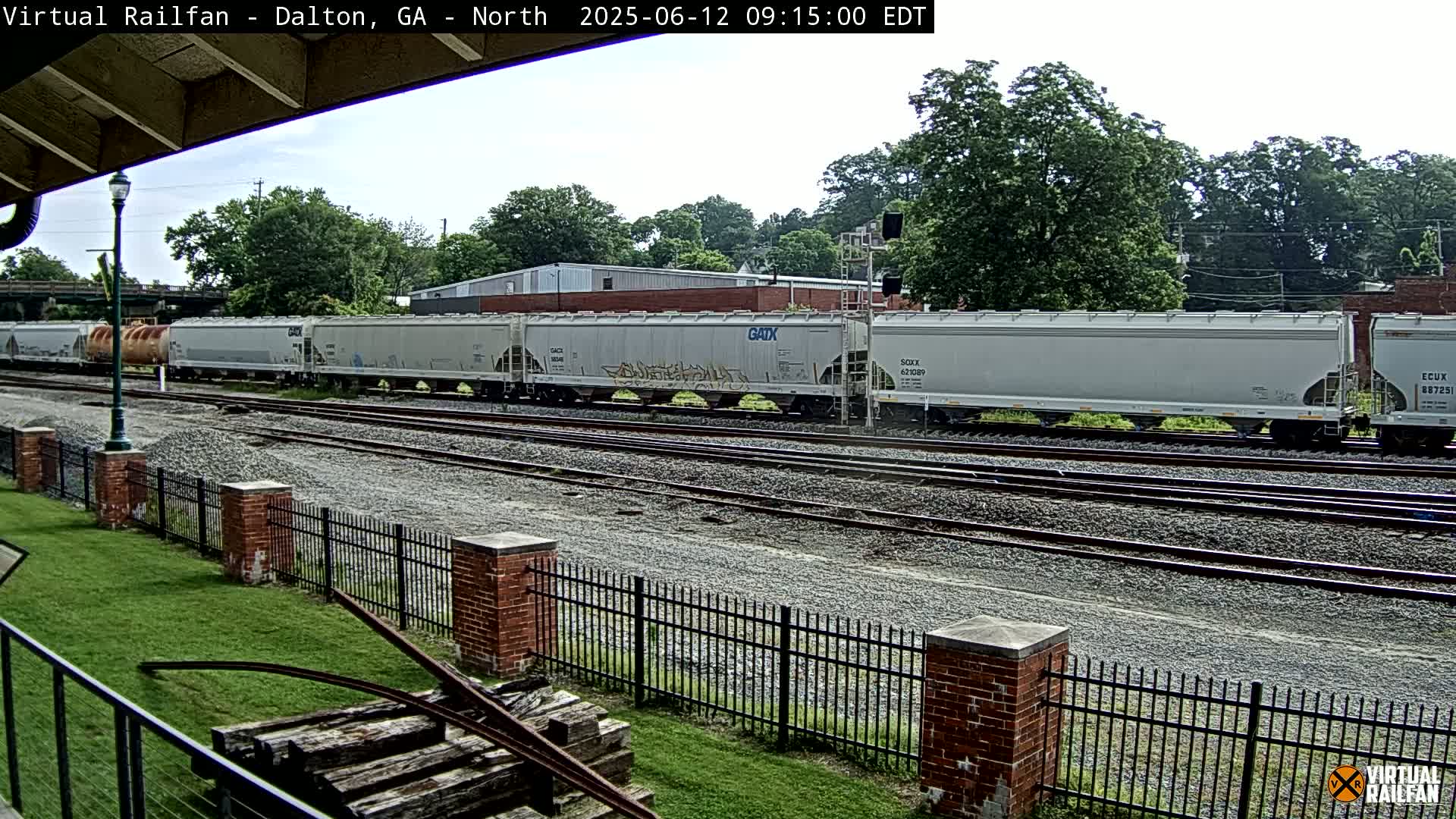 A long freight train of light gray covered hopper cars sits on a rail line next to a grassy area with a brick fence, under a partly cloudy sky.