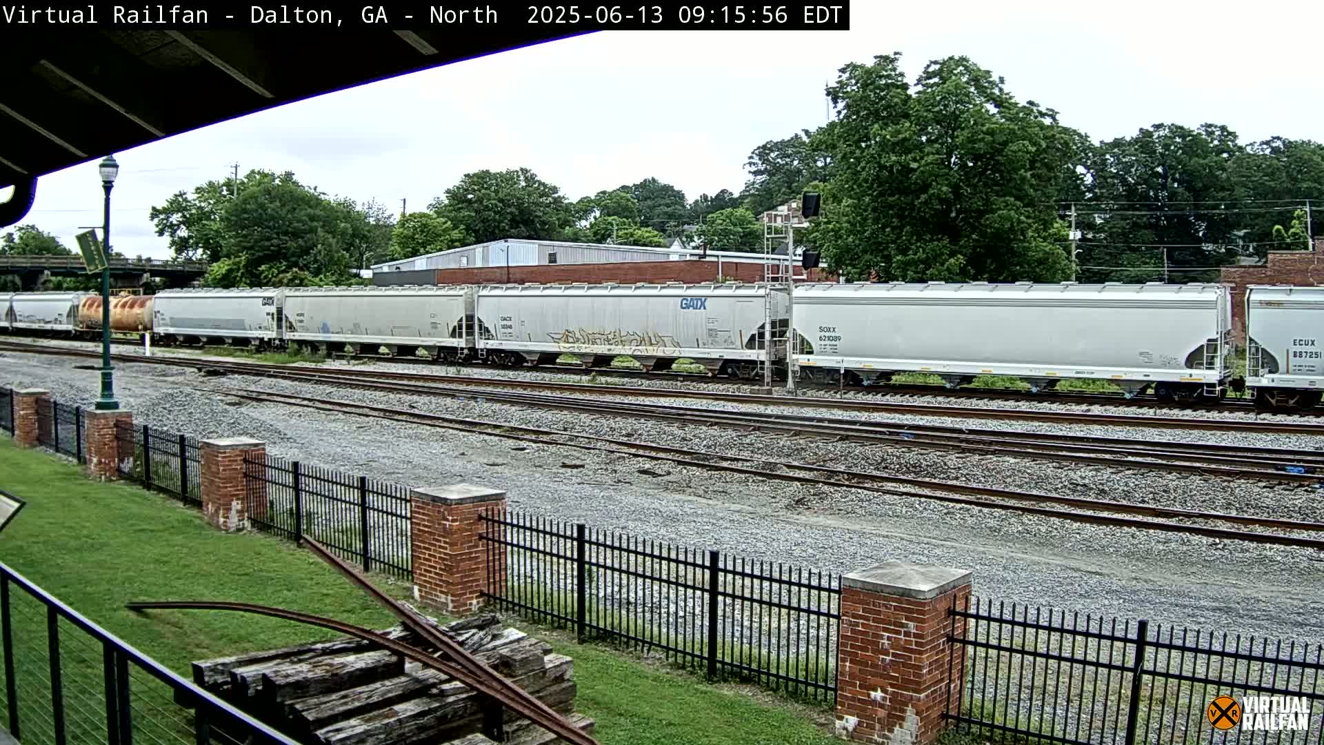 A long freight train of light gray covered hopper cars sits on a rail line next to a brick fence and grassy area, under an overcast sky.