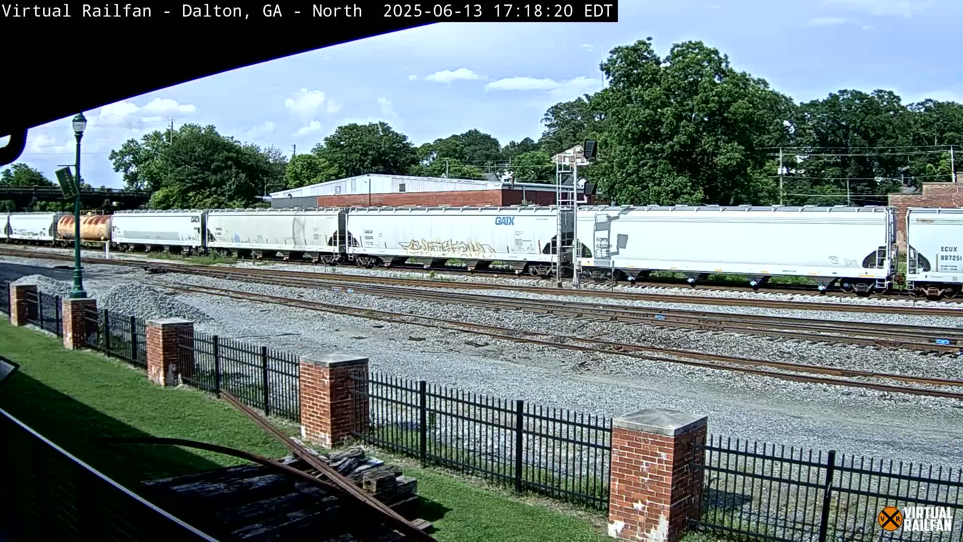 A long freight train of mostly white boxcars, some with graffiti, sits on a track next to a brick fence under a partly sunny sky.
