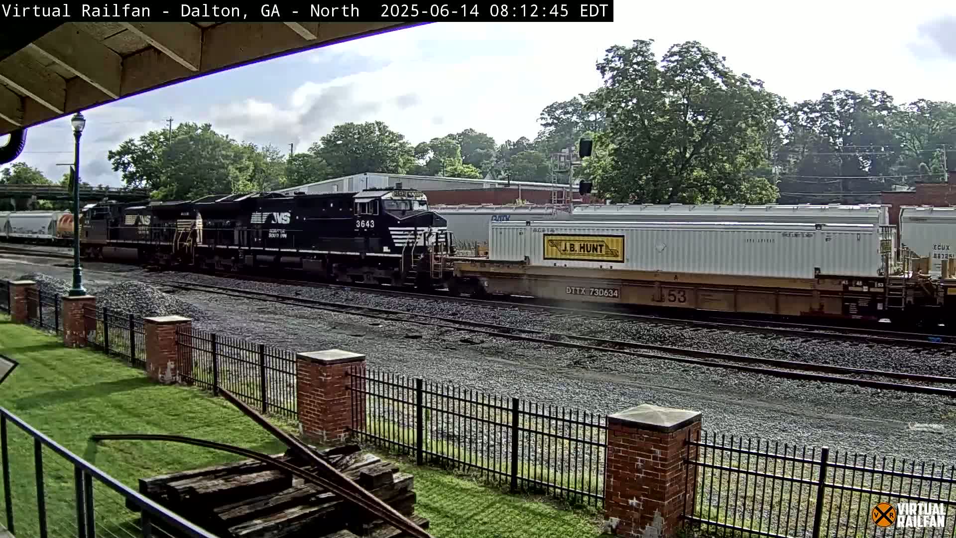A black diesel locomotive leads a freight train past a brick fence under partly cloudy skies.