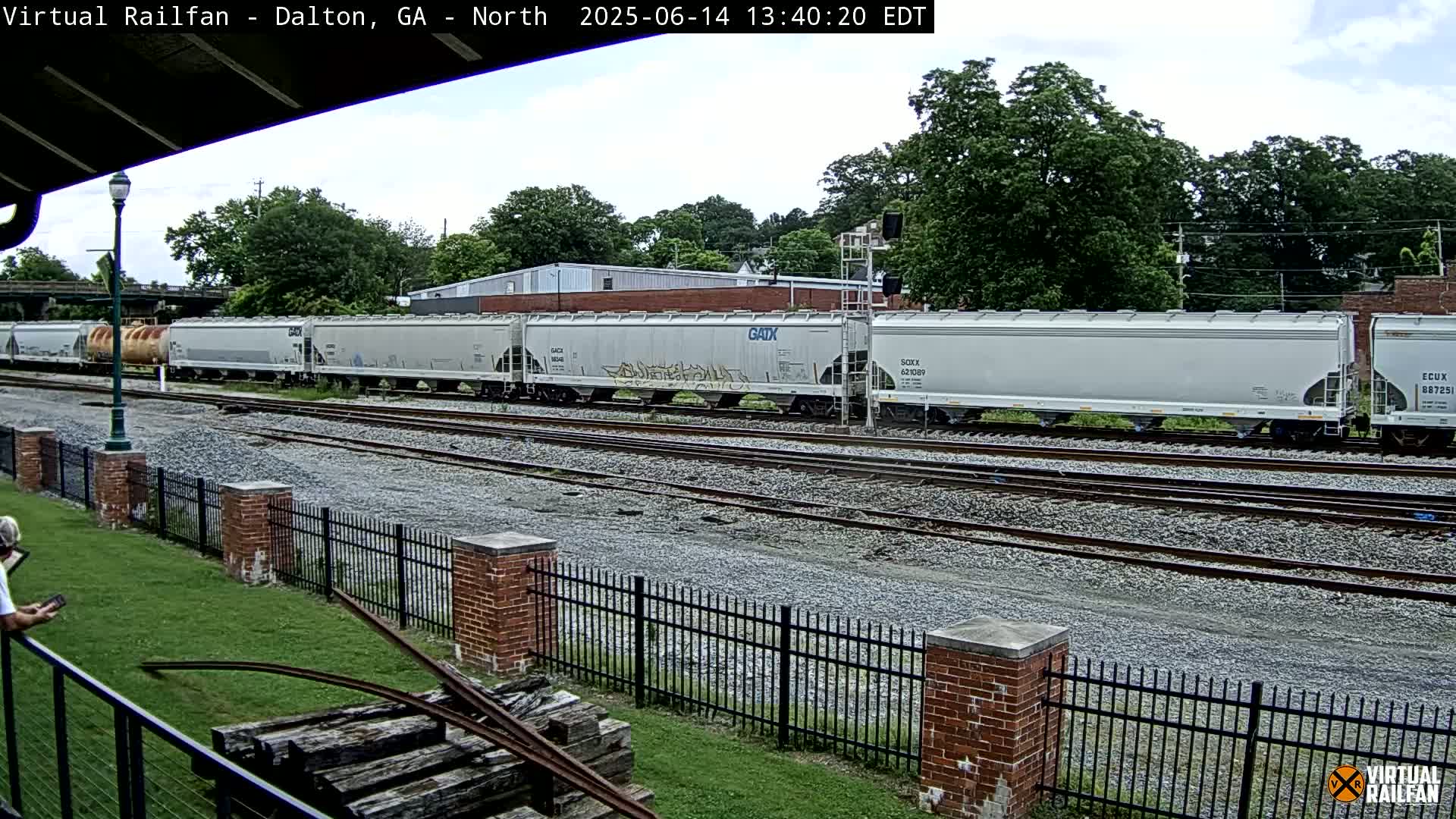 A long freight train of light gray covered hopper cars sits on a rail line next to a grassy area with a brick fence under a partly cloudy sky.