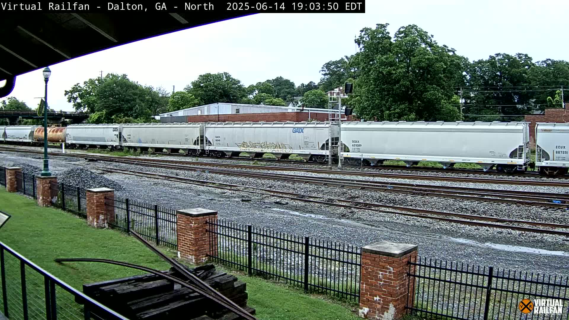 A long freight train of light gray covered hopper cars sits on a rail line next to a grassy area with a brick fence under a partly cloudy sky.