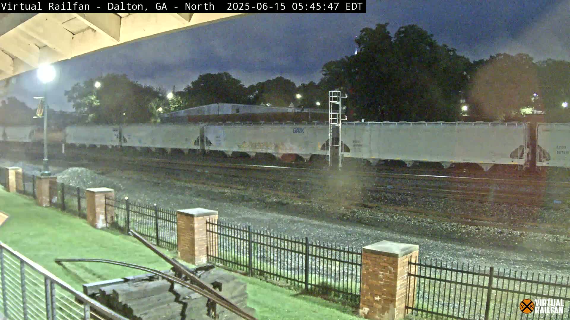 A long freight train of light-grey cars sits on railroad tracks under a dark, possibly overcast sky at night.