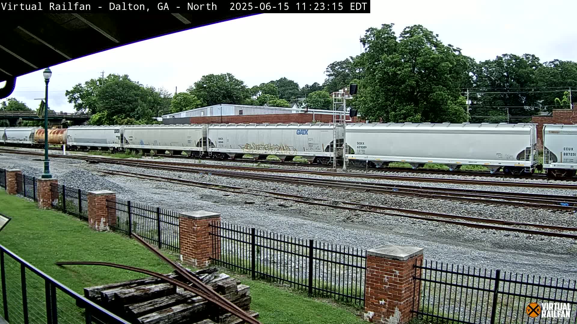 A long freight train of light gray covered hopper cars sits on a set of railroad tracks next to a grassy area with a brick fence under an overcast sky.