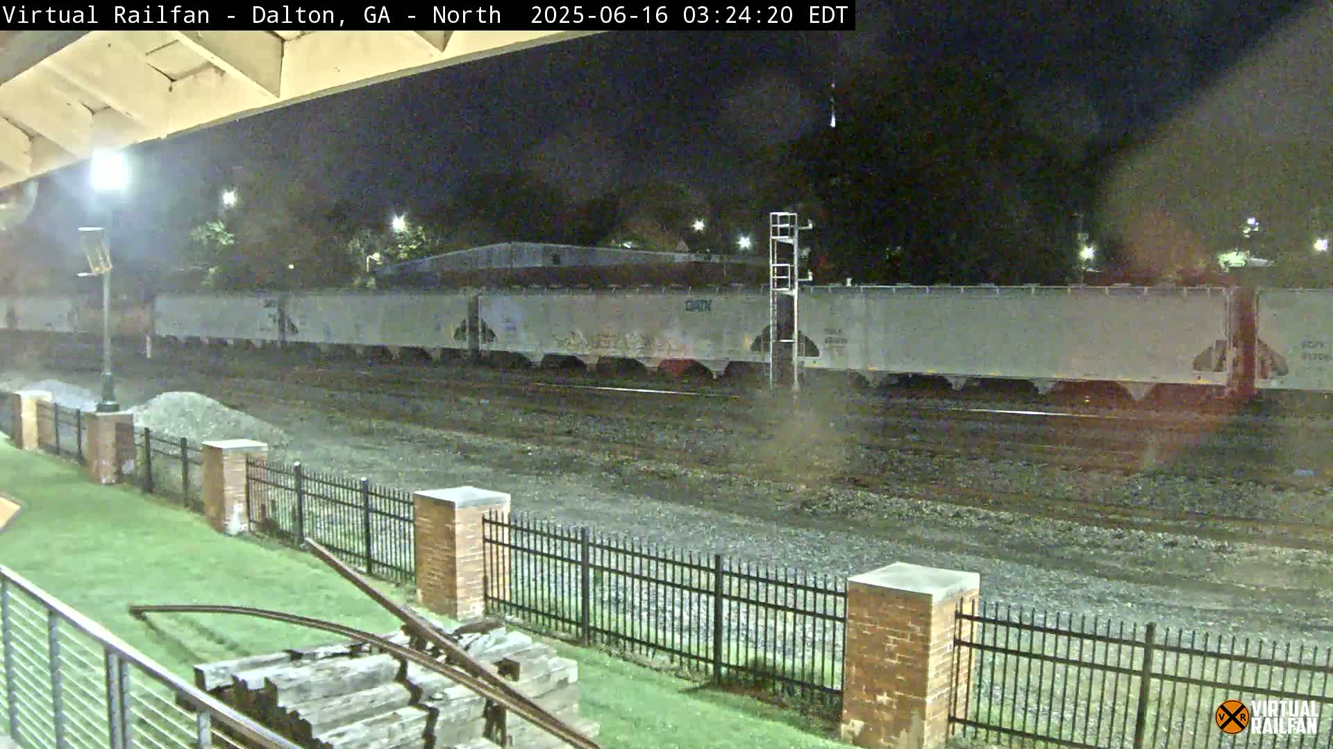 At night, a long freight train of light gray boxcars sits on railway tracks next to a grassy area enclosed by a black metal fence and brick pillars.