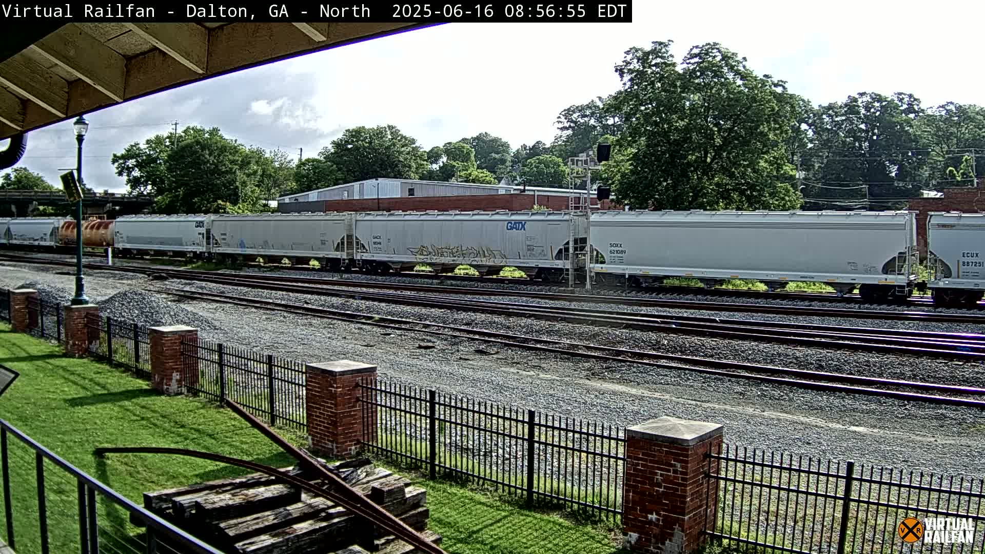 A long freight train of light gray covered hopper cars sits on a set of train tracks next to a grassy area with a brick fence under a partly cloudy sky.