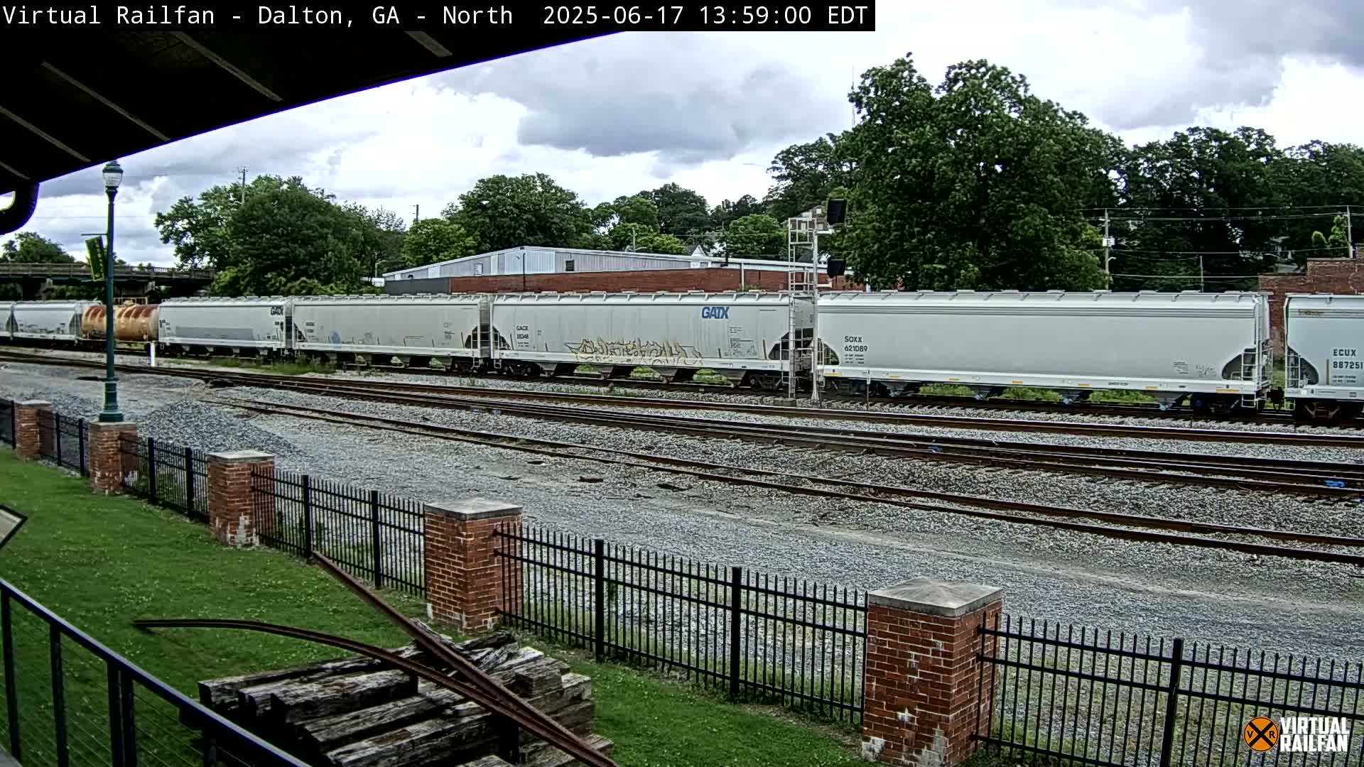 A long freight train of light gray covered hopper cars sits on a rail line under a partly cloudy sky, next to a grassy area with a brick fence.