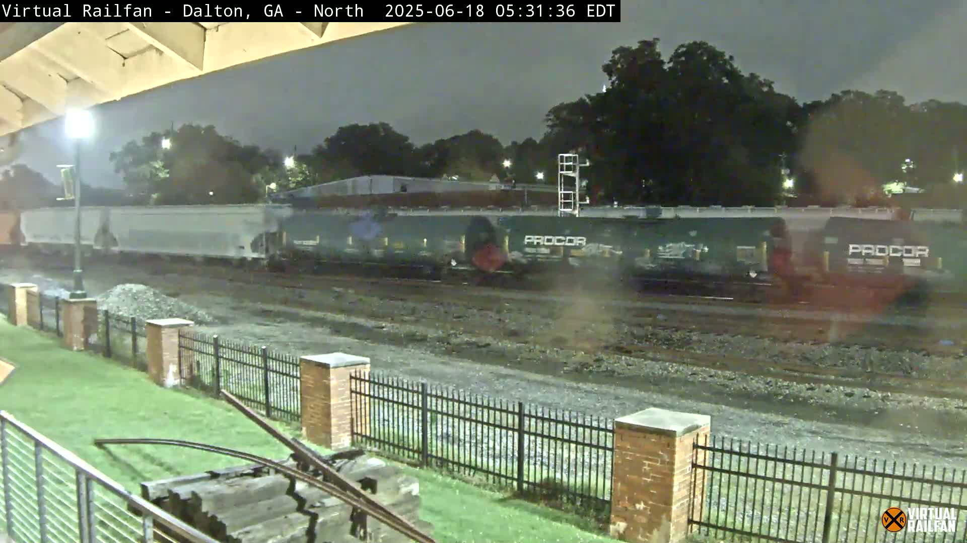 A long freight train passes by at night, under a dark sky, next to a grassy area with a brick fence and some railroad infrastructure.