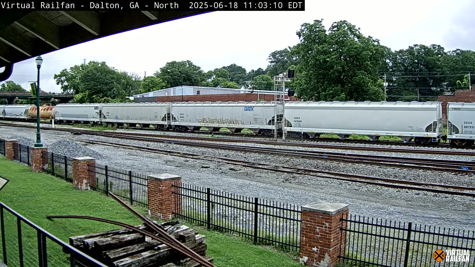 A long freight train of light gray boxcars sits on a set of railway tracks under an overcast sky.