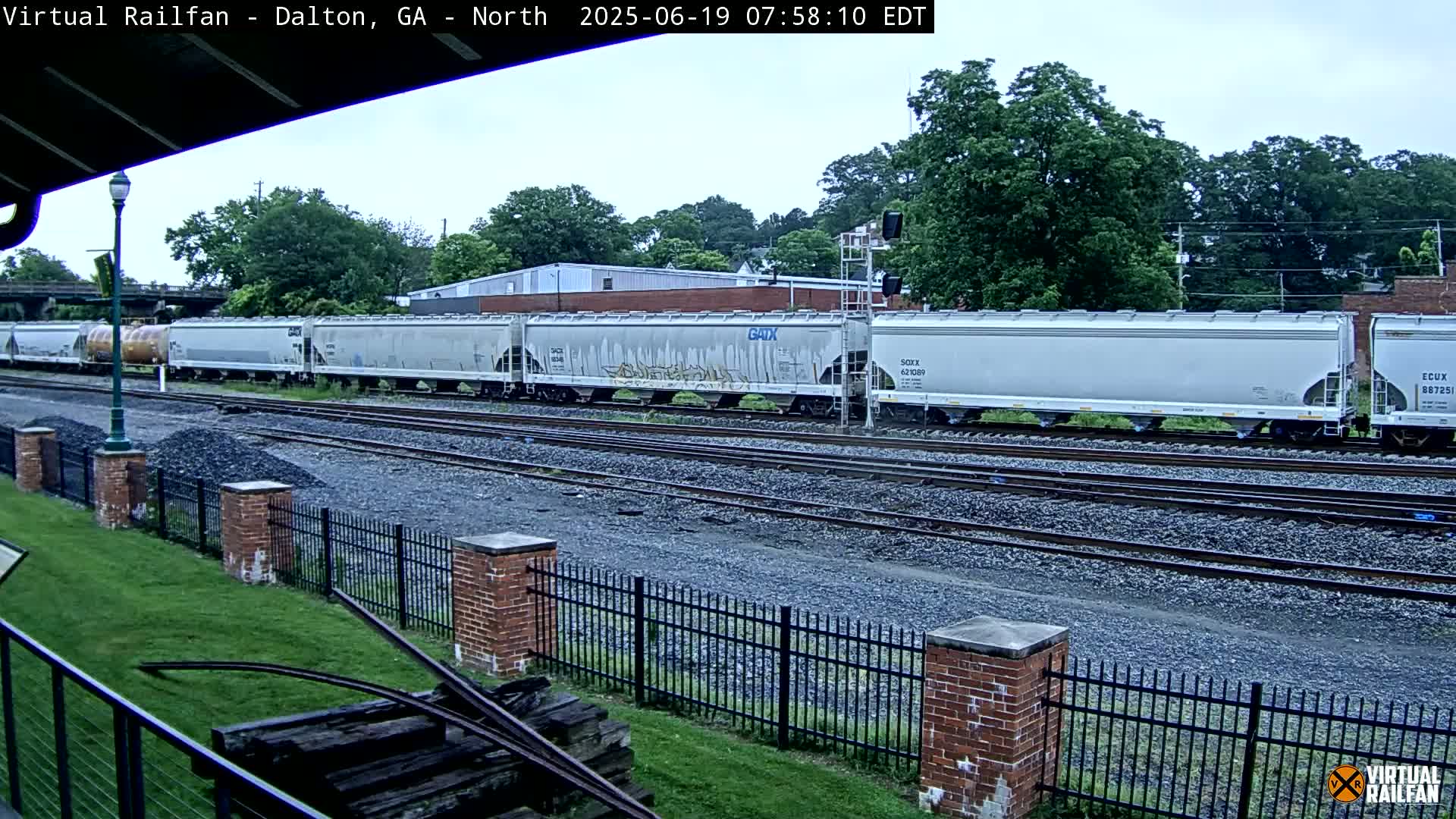 A long freight train of covered hopper cars passes by on a cloudy day next to a grassy area with a brick fence and some old railway equipment.