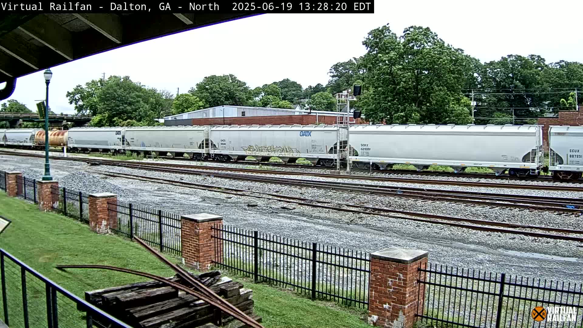 A long freight train of light gray covered hopper cars sits on a railway track under an overcast sky, beyond a brick fence and grassy area.
