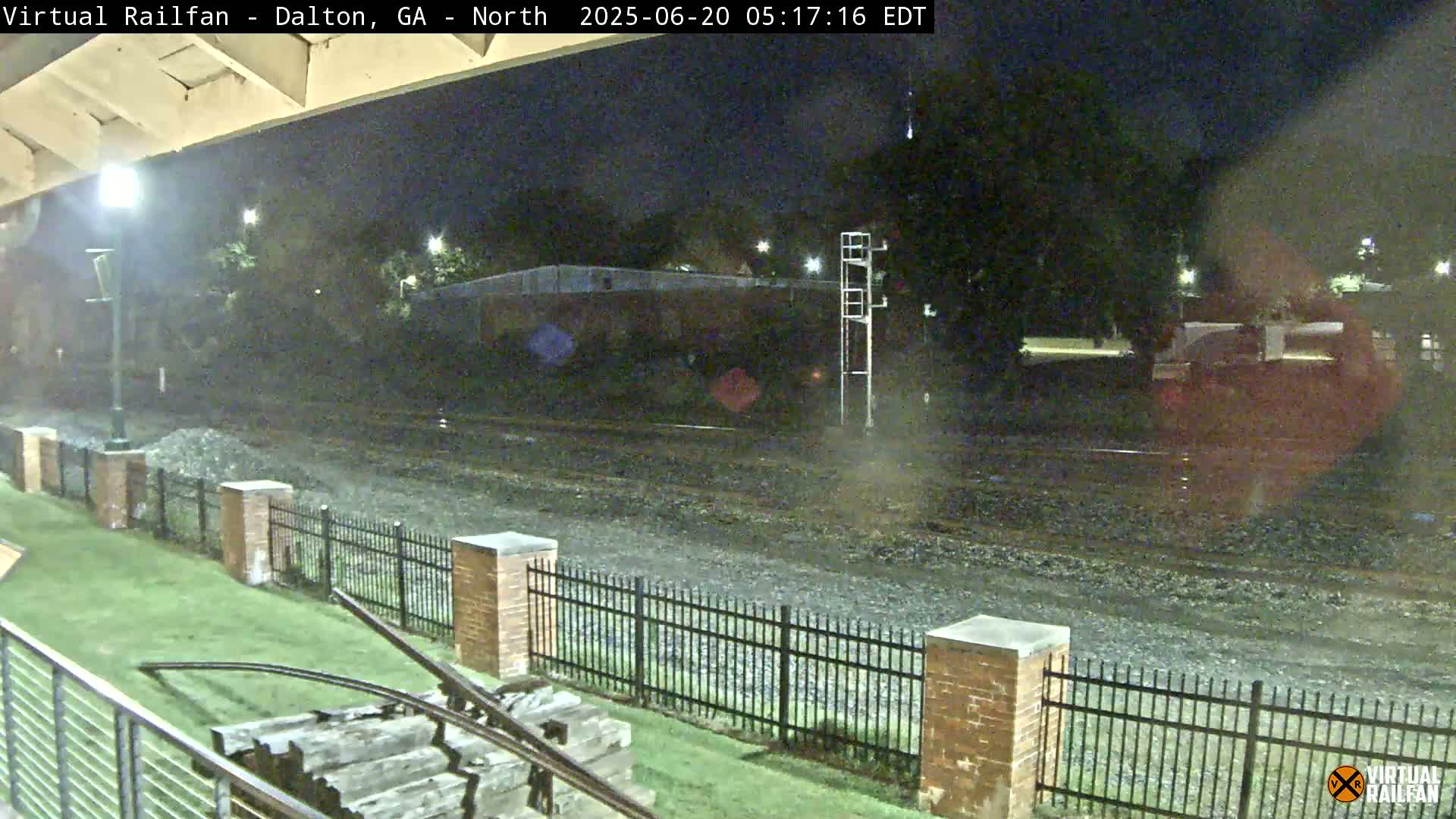A nighttime view of railroad tracks and a grassy area with a metal fence, under a dark sky.