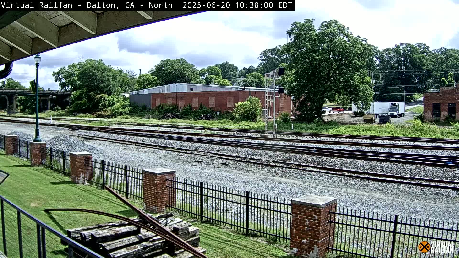 A partly cloudy day reveals multiple train tracks bordered by a brick fence and green grass, with industrial buildings and trees in the background.