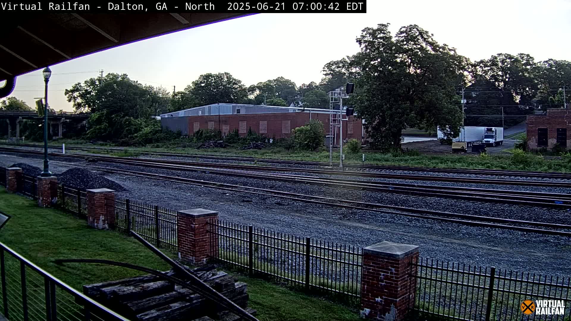 Multiple train tracks stretch into the distance under a partly cloudy morning sky, bordered by a brick fence and lush green grass.