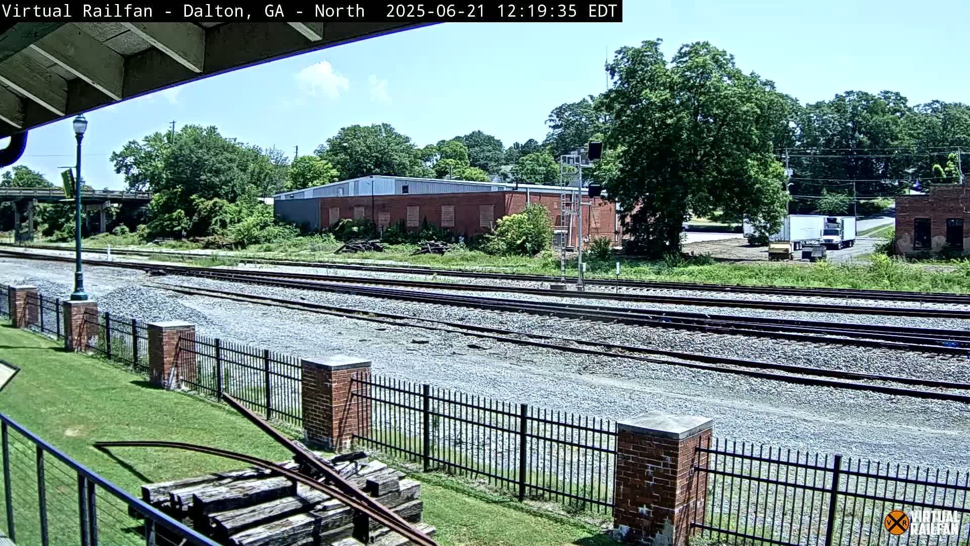 Multiple train tracks stretch into the distance under a partly sunny sky, bordered by a brick fence, a grassy area, and several buildings.