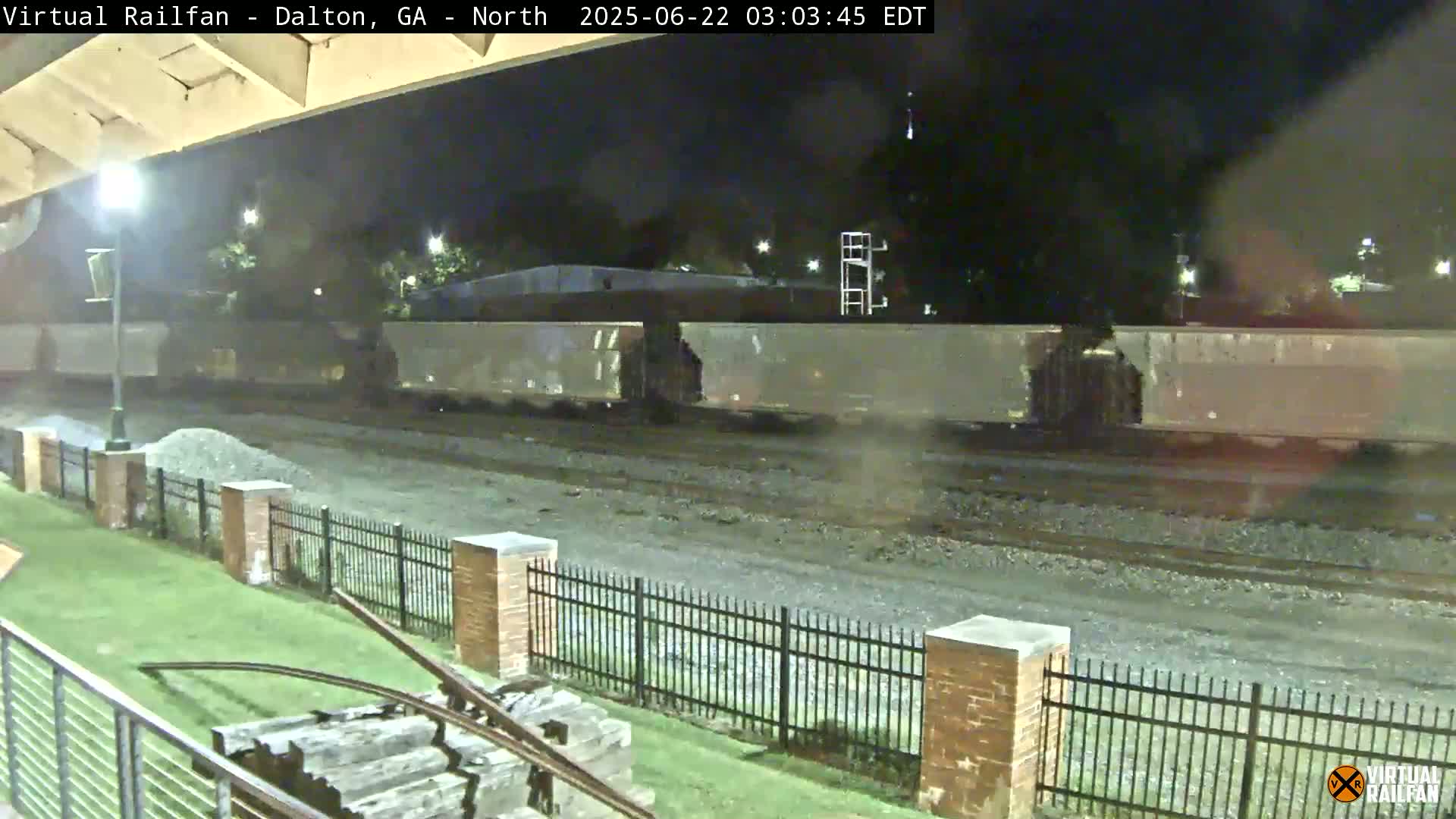 A nighttime view of a long freight train passing by a fenced area with a grassy lawn and some old railway equipment visible in the foreground.