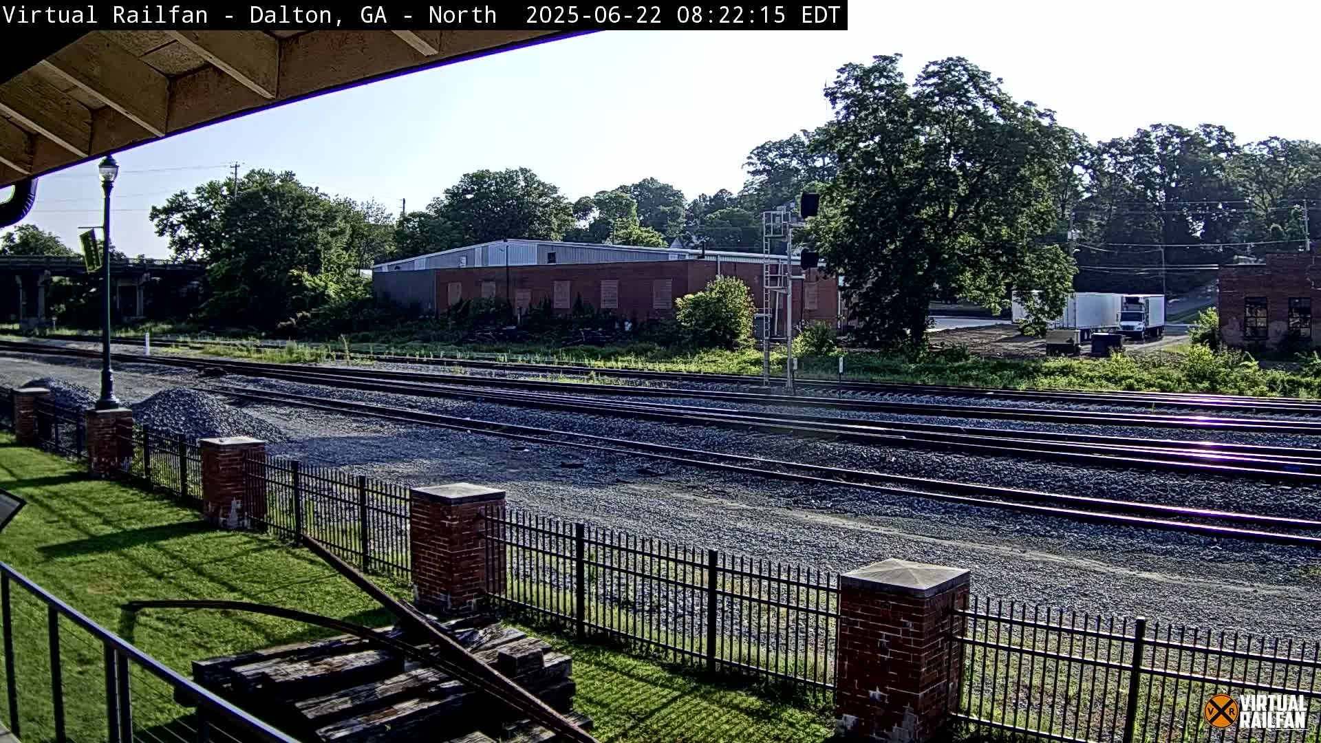 A sunny view of multiple train tracks stretches from a grassy area with a brick fence, towards buildings and trees in the distance.