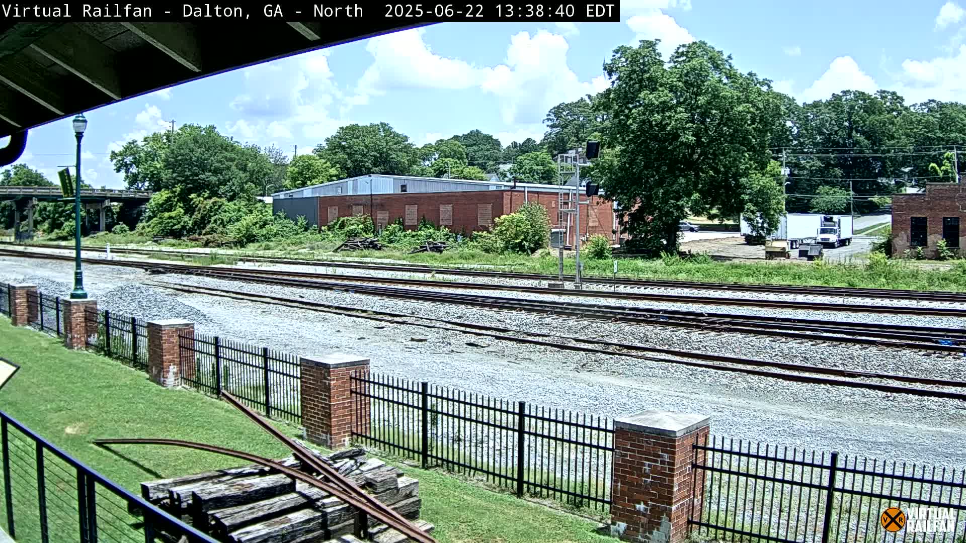 Multiple train tracks run alongside a brick fence, separating a grassy area from a cluster of buildings and lush green trees under a partly cloudy sky.