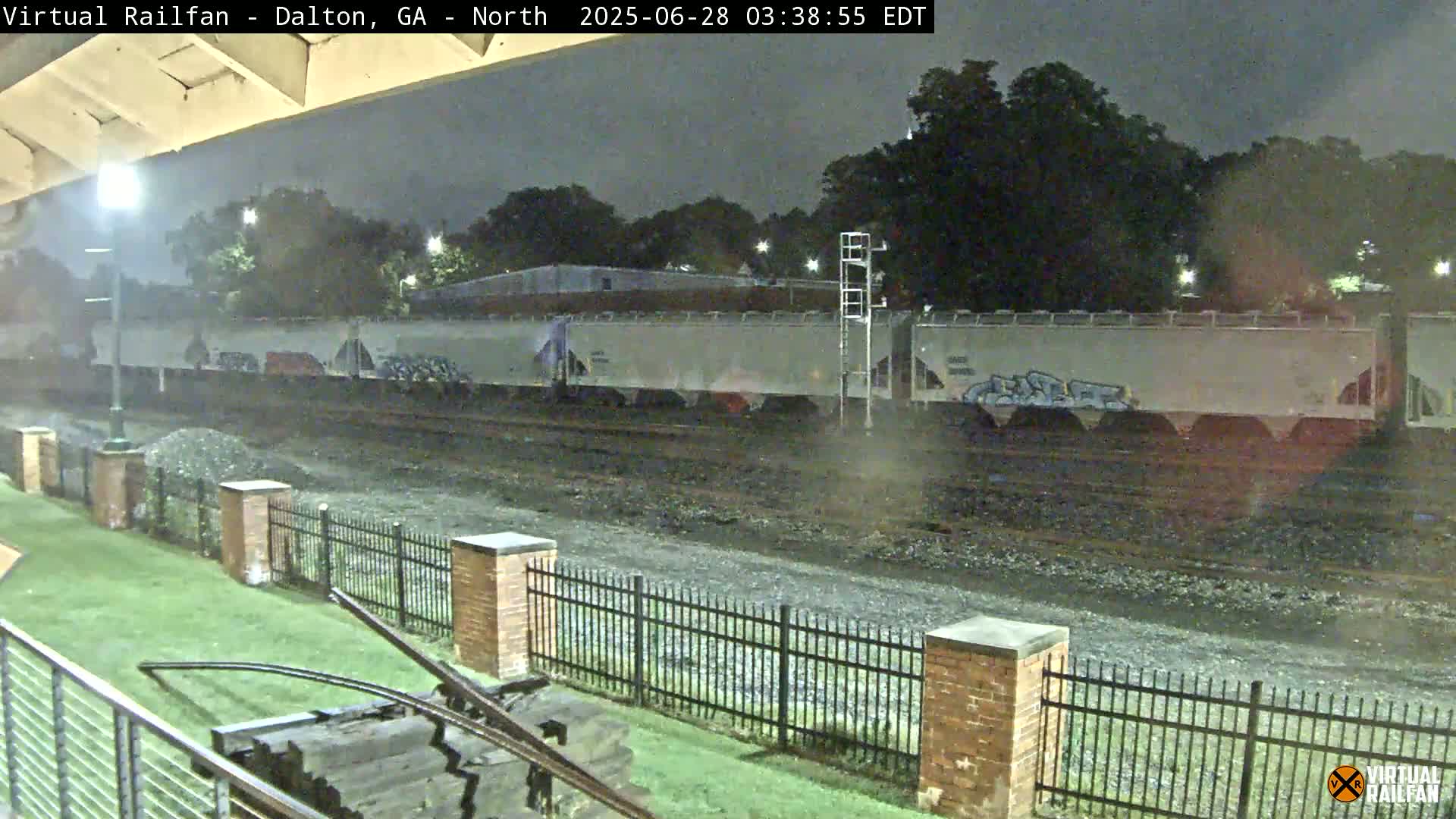 At night, a long freight train sits on railway tracks next to a grassy area enclosed by a black metal fence and brick pillars.