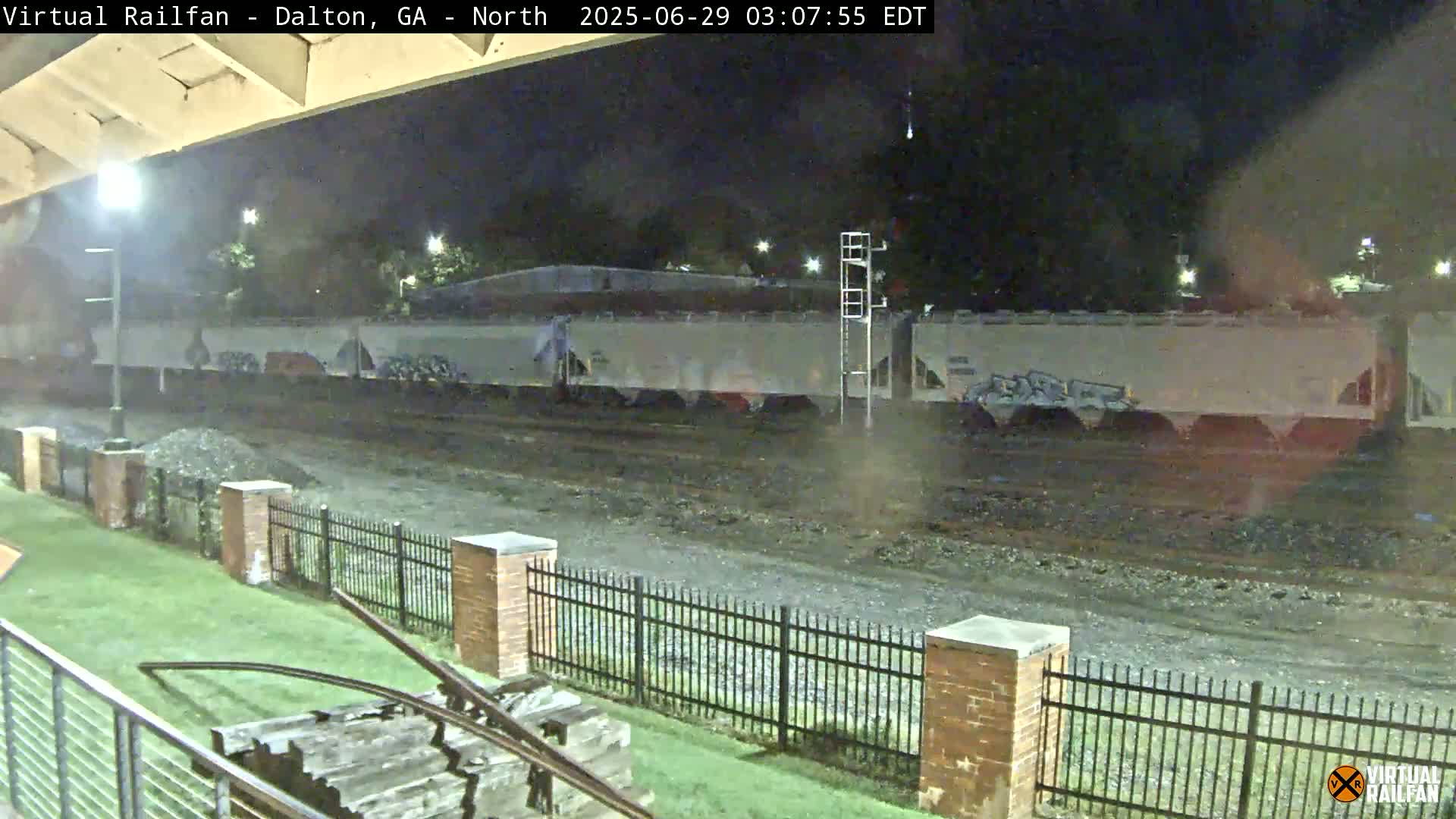 At night, a long freight train sits on tracks behind a short black metal fence, with a grassy area and brick pillars in the foreground.