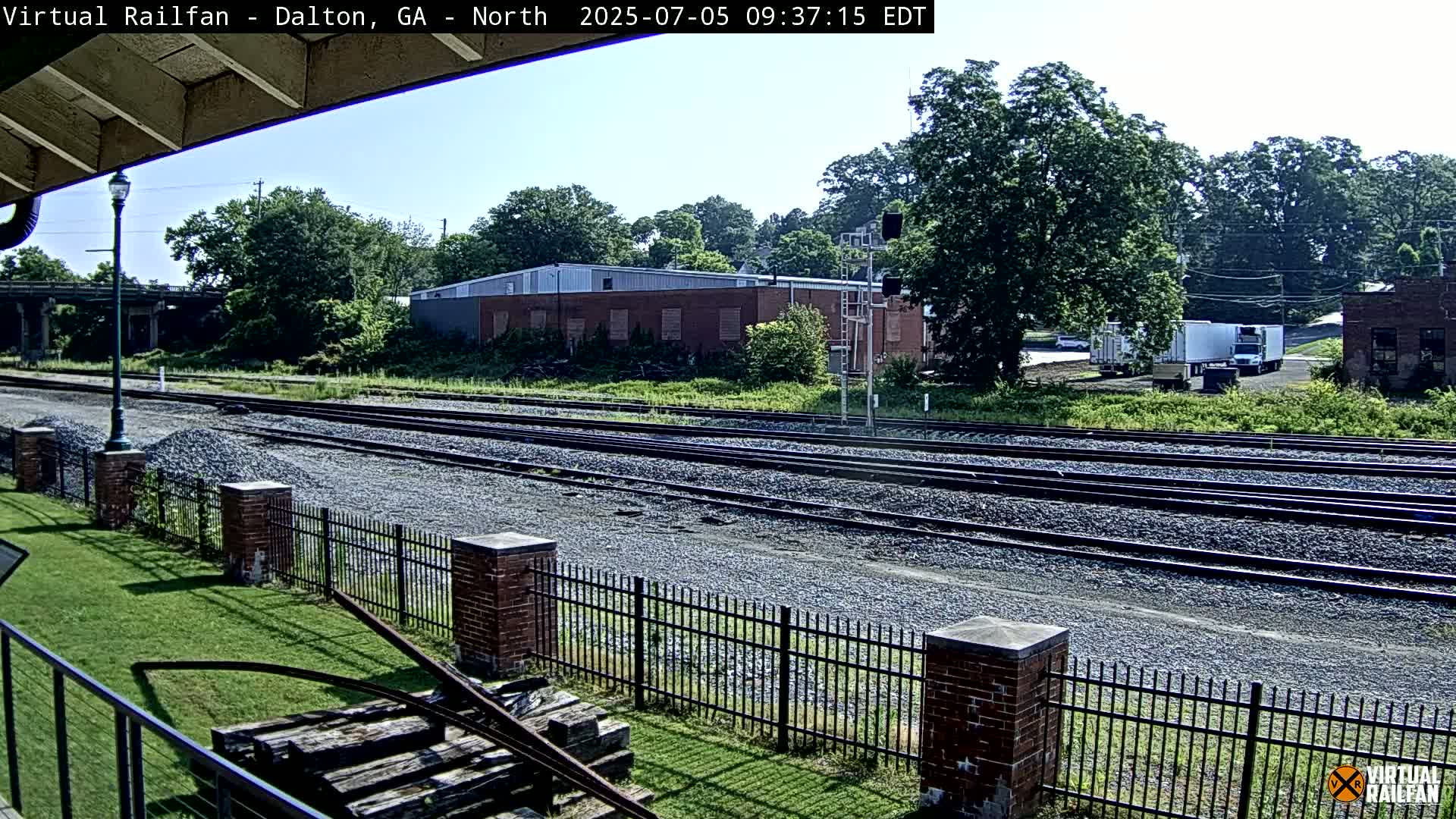 Multiple train tracks stretch into the distance past a brick fence and grassy area, under a clear sky and beside industrial buildings and trees.