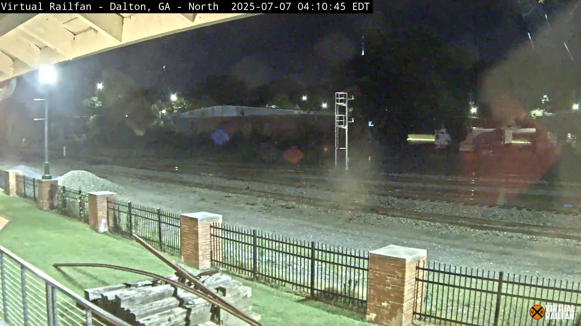 A nighttime view of train tracks behind a brick fence, with some grass and a piece of old railway equipment in the foreground, under a slightly overcast sky.