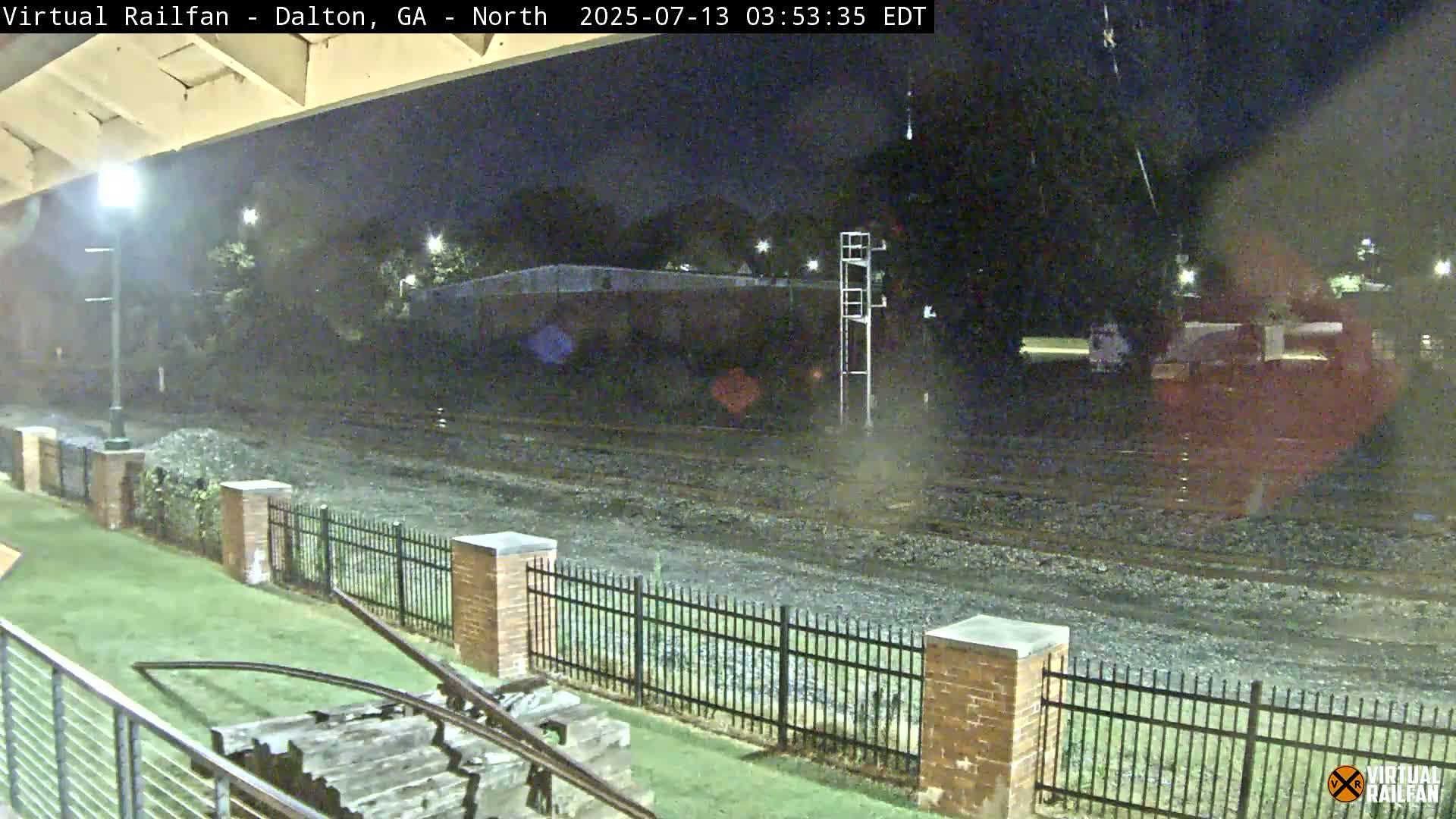 A nighttime view of train tracks, bordered by a metal fence and brick pillars, with a grassy area and part of a structure visible in the foreground.