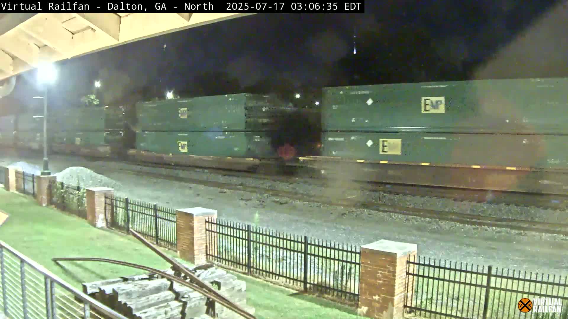 At night, a long freight train carrying stacked green shipping containers passes by a brick fence and grassy area.
