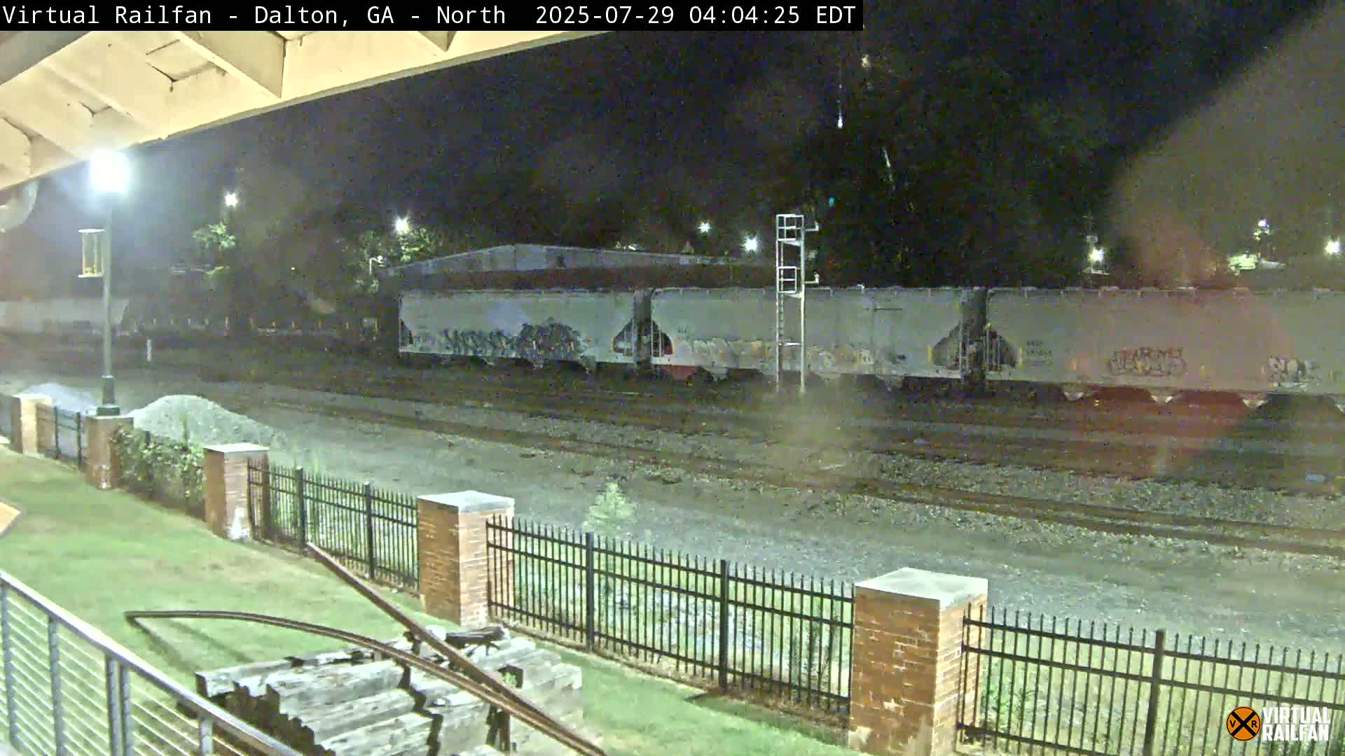 At night, a freight train with graffiti-covered cars sits on the tracks next to a grassy area with a brick fence.