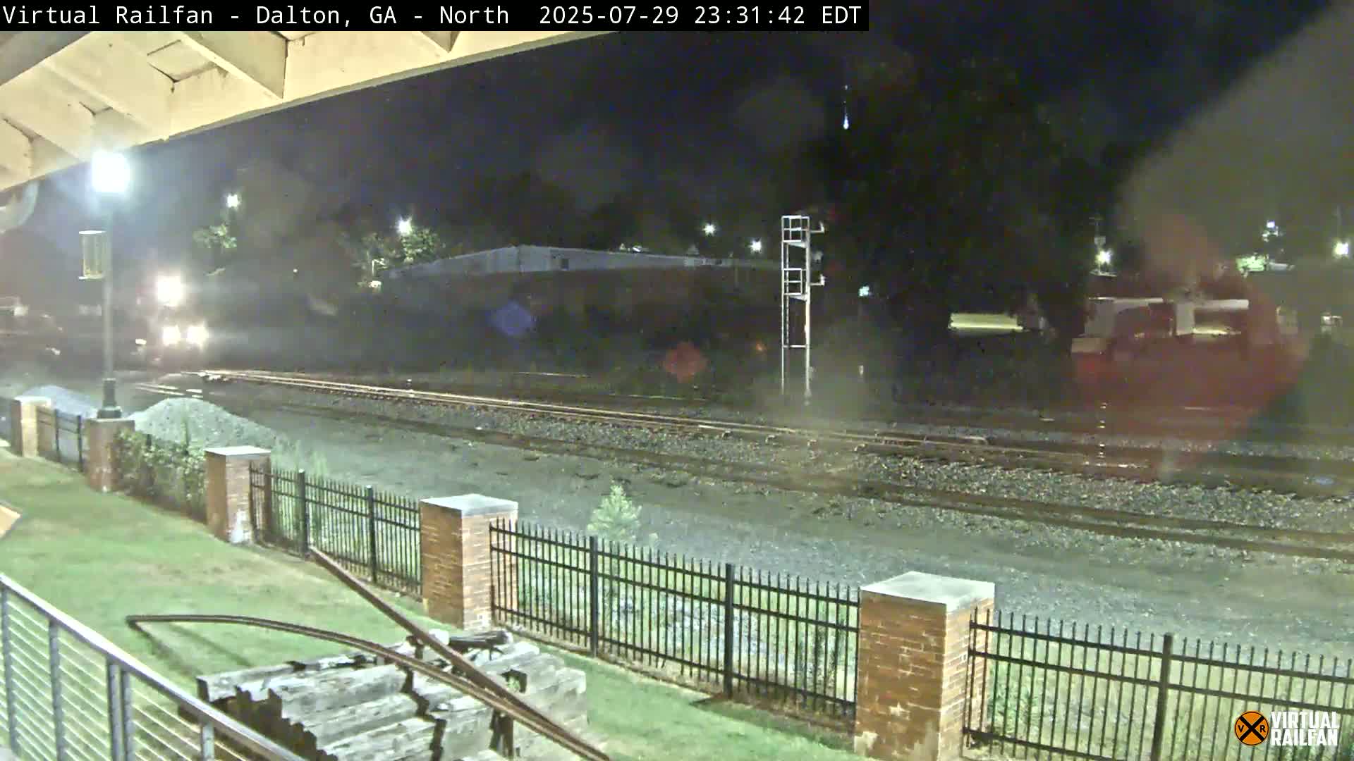 At night, a freight train with graffiti-covered cars sits on the tracks next to a grassy area with a brick fence.