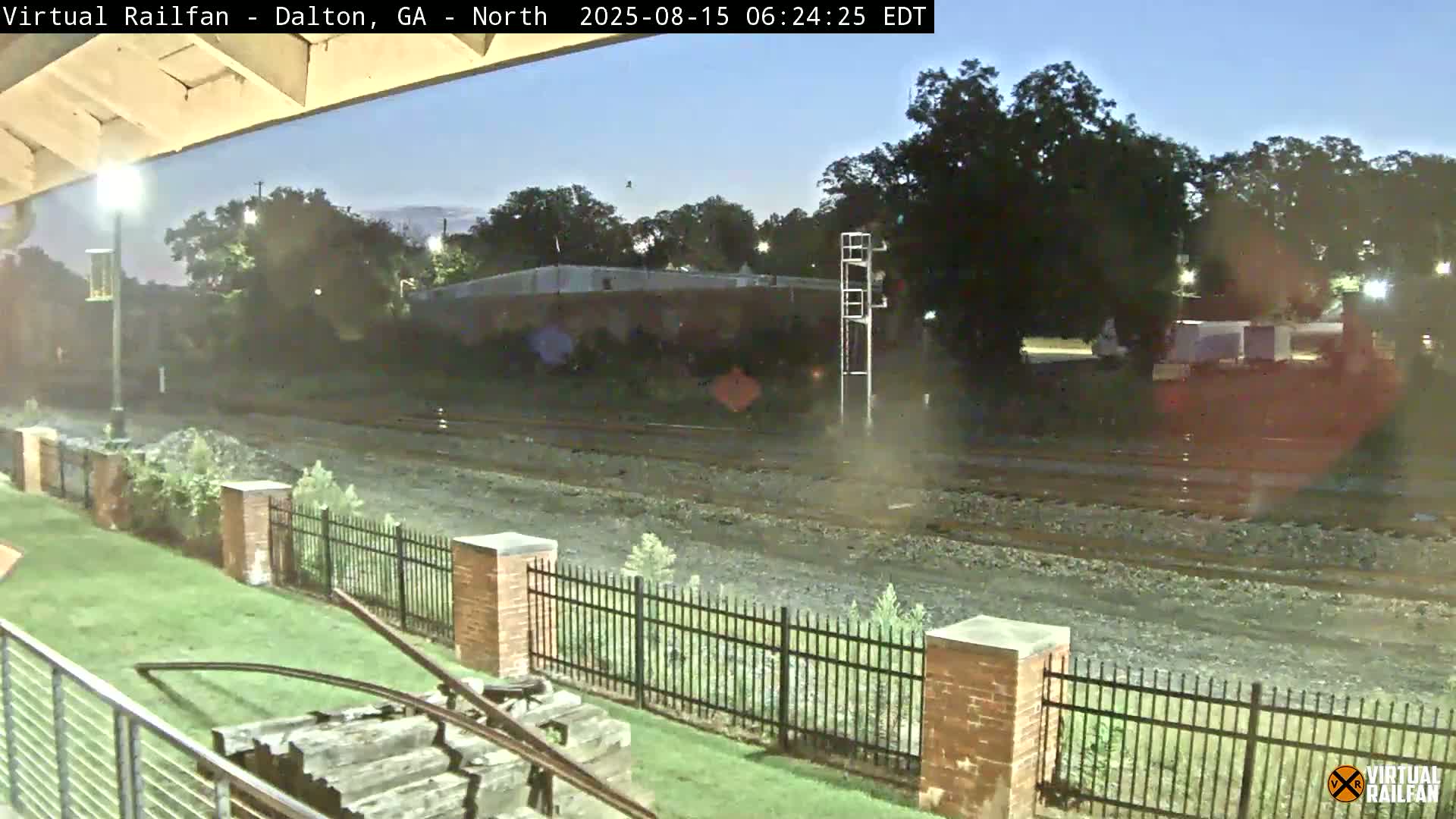 At night, a freight train with graffiti-covered cars sits on the tracks next to a grassy area with a brick fence.