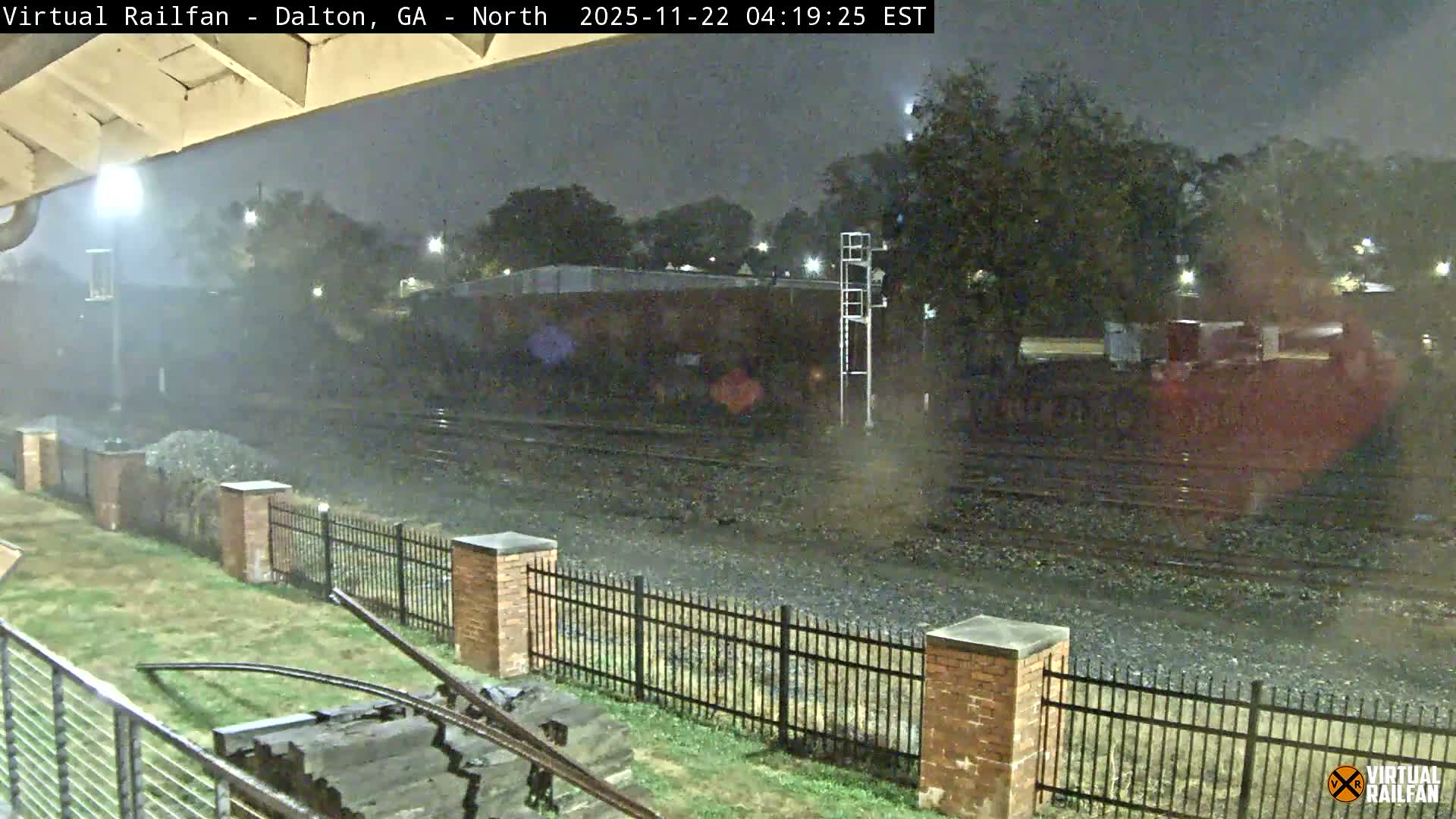 A nighttime scene captures several wet railroad tracks in the rain, flanked by a metal fence and grassy area in the foreground, with distant buildings and trees faintly visible under the glow of streetlights.