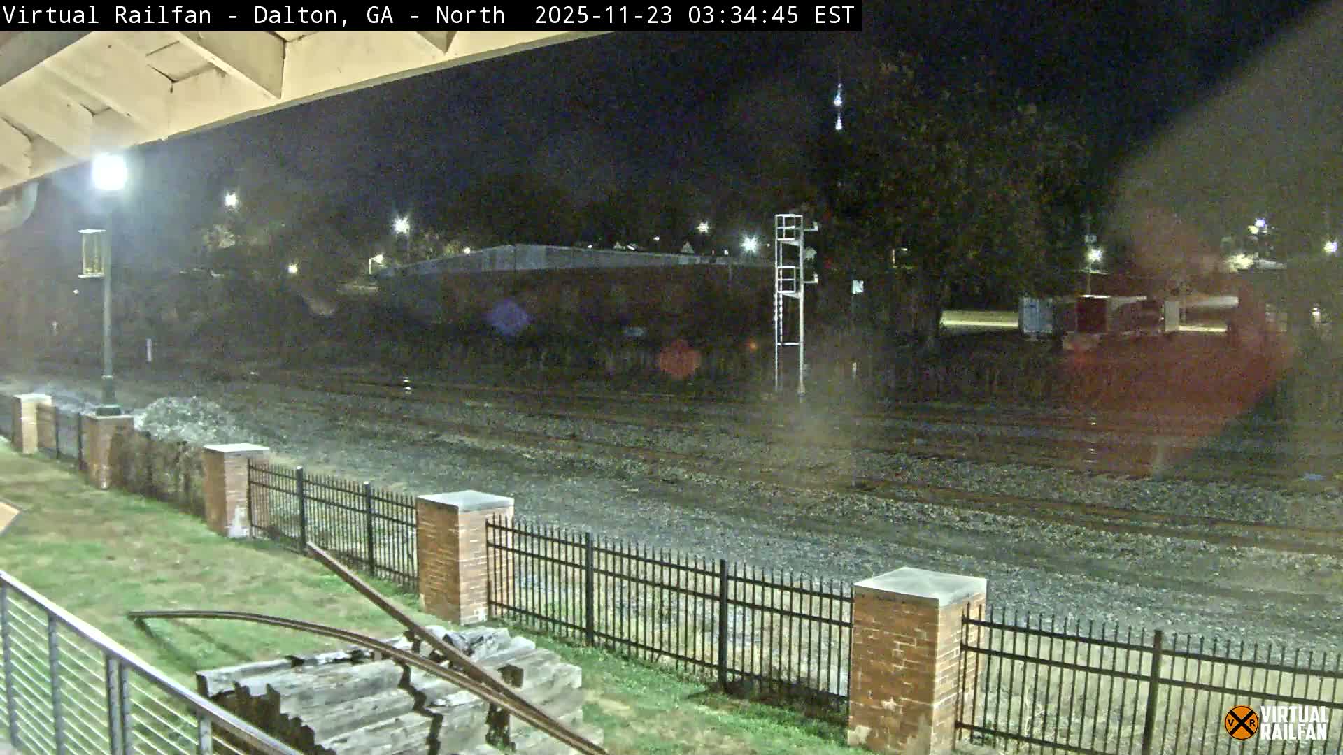 Multiple railroad tracks are seen stretching into the distance on a damp night, illuminated by numerous streetlights, with a grassy area and a black metal fence running parallel to the tracks in the foreground, viewed from beneath an overpass.