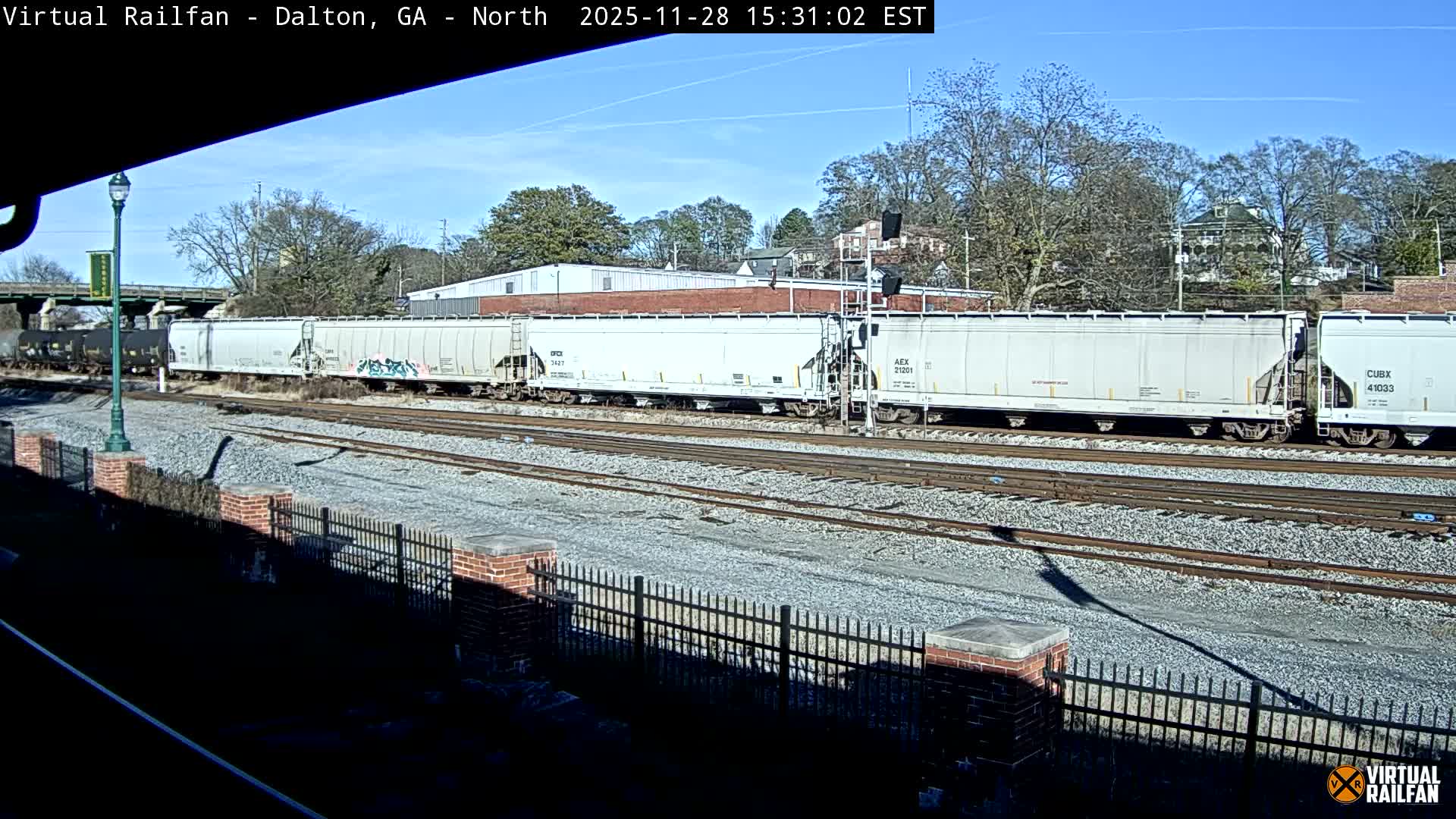 A long freight train, featuring white hopper cars and black tanker cars, rests on multiple tracks under a clear, sunny sky with distant buildings and bare trees in the background.