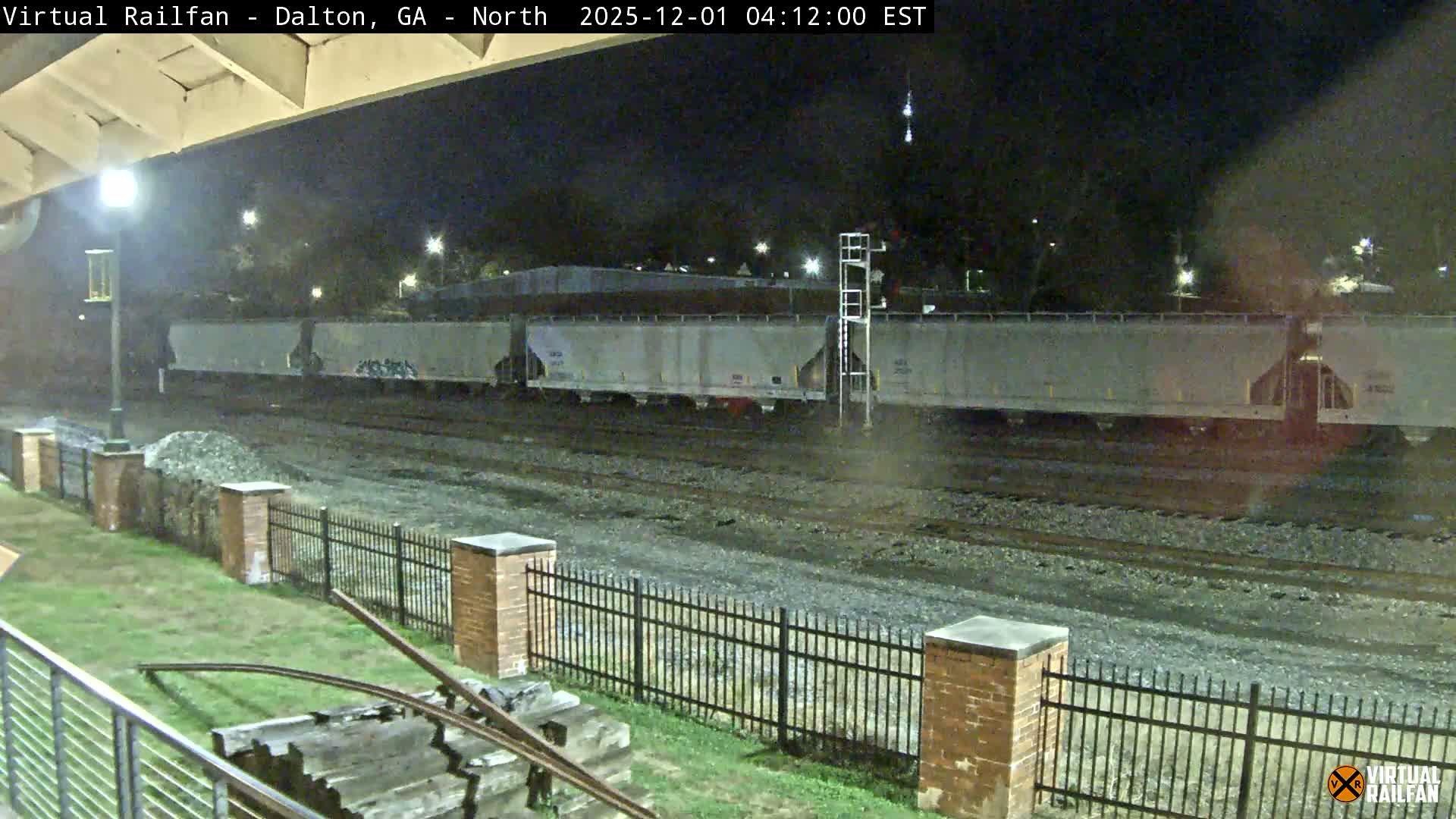 A long freight train rests on multiple tracks at night under a clear sky, with a grassy area bordered by a black metal fence and brick posts in the foreground.