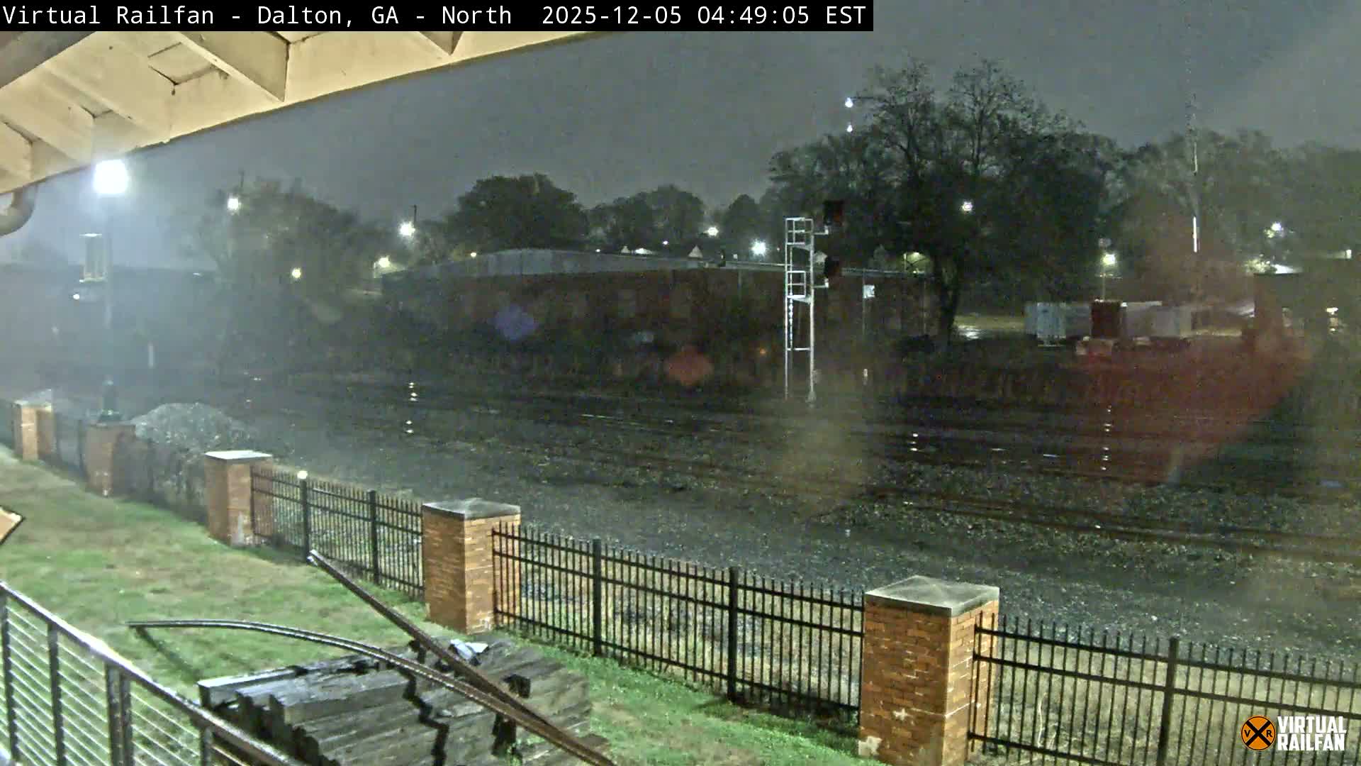 The image captures a dark, rainy night at a railway yard, with multiple tracks glistening wet, a signal tower standing tall, and a black metal fence bordering the foreground, all under the blur of falling rain illuminated by scattered lights.