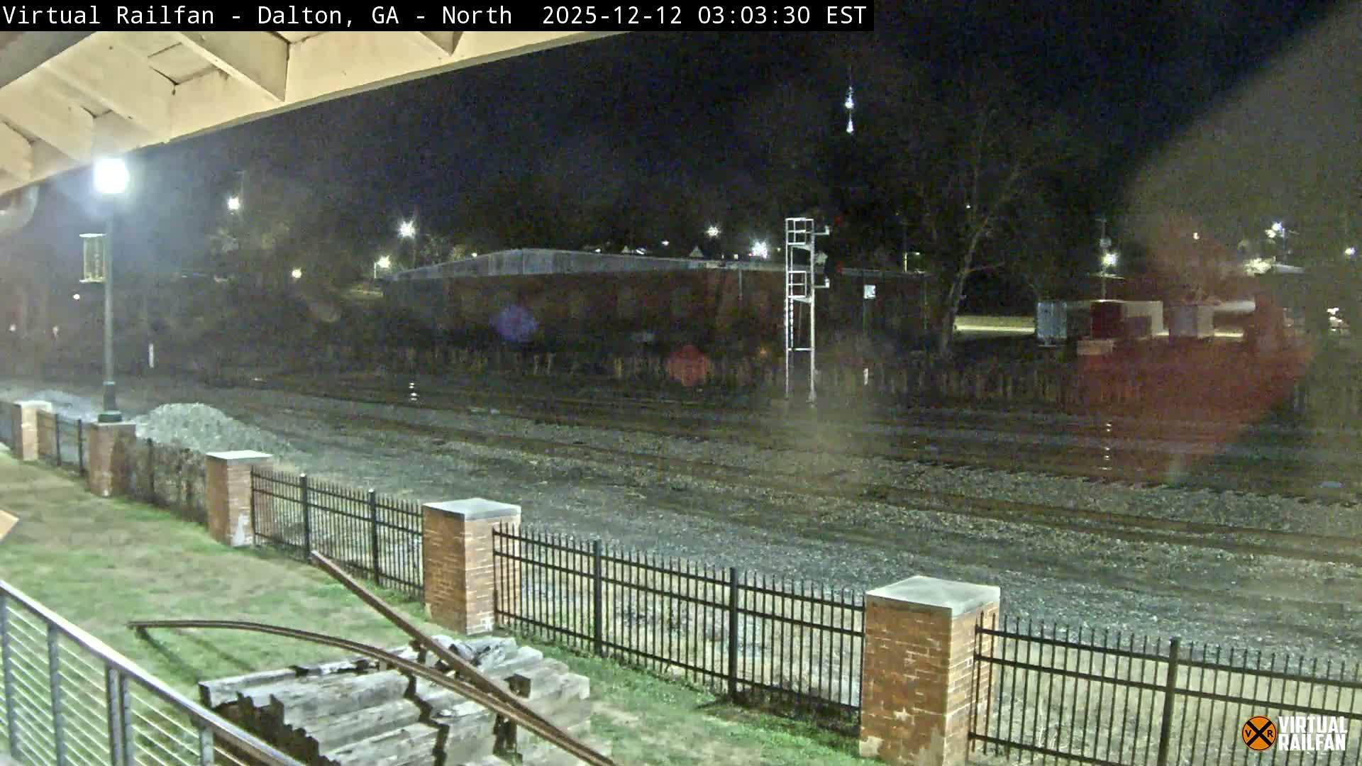 A rain-slicked train yard is seen at night, featuring multiple tracks alongside a grassy area with a black fence and discarded railway ties in the foreground, illuminated by distant industrial lights and streetlights.