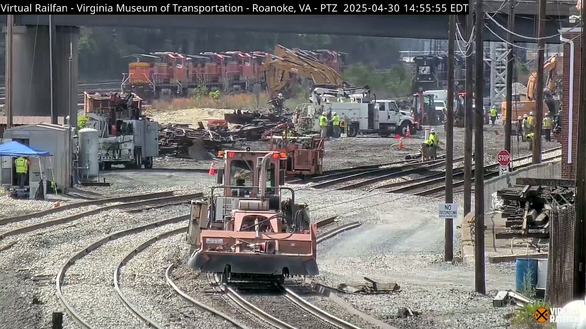 A sunny day at a railroad yard shows several workers and pieces of heavy machinery near damaged train cars and rail lines.