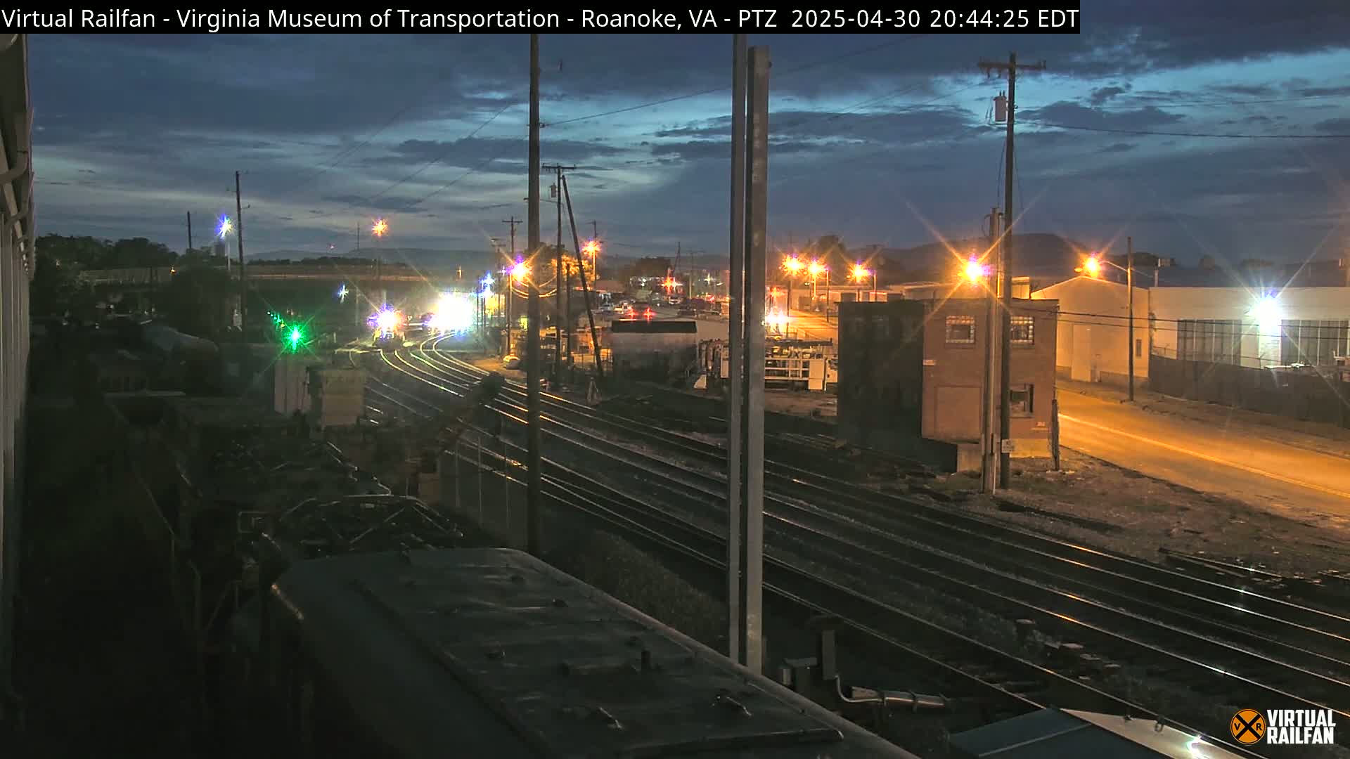 A nighttime view of railroad tracks and train cars, under a partly cloudy sky, with streetlights illuminating nearby buildings and a road.