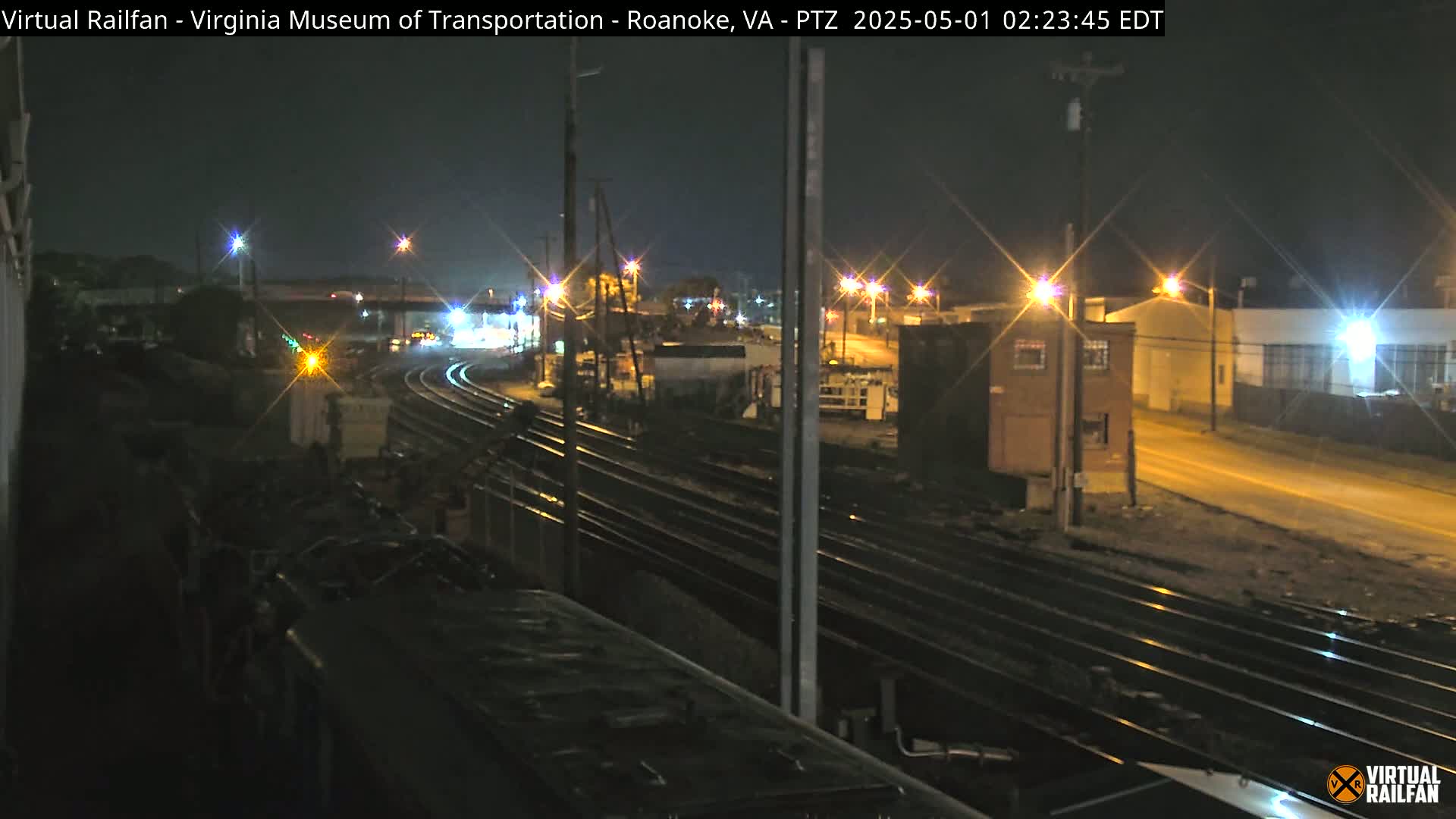 At night, a train sits near several sets of train tracks that curve around industrial buildings and a road with streetlights.