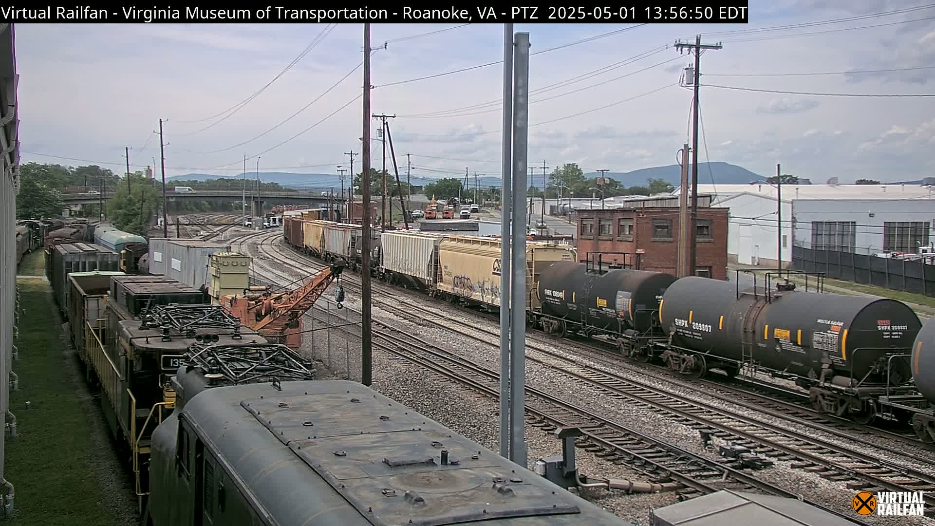 A long freight train with various cargo cars and locomotives sits on a railway track under an overcast sky, with other trains and industrial buildings visible in the distance.