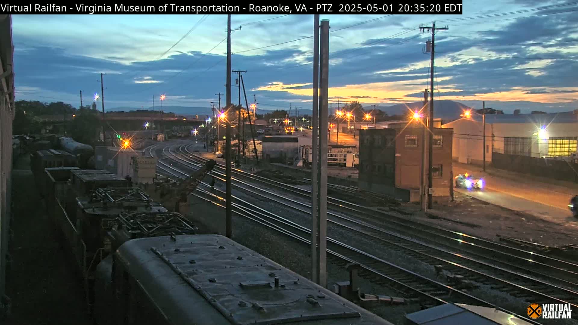 A twilight view of railroad tracks and yards, with several trains and buildings visible under a partly cloudy sky.
