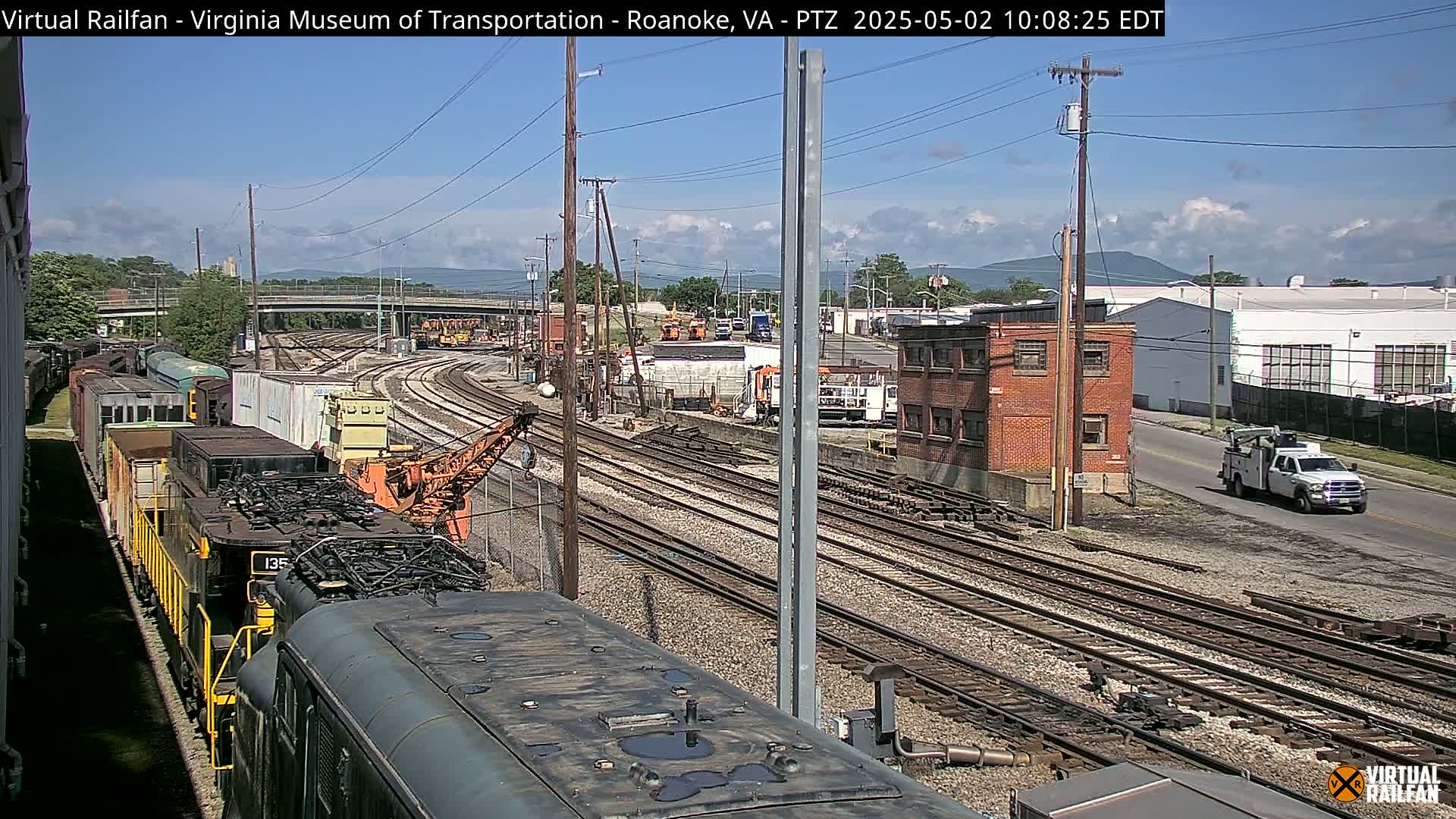 A mostly sunny day shows a train yard with multiple rail lines, a locomotive, several freight cars, and industrial buildings in the background.