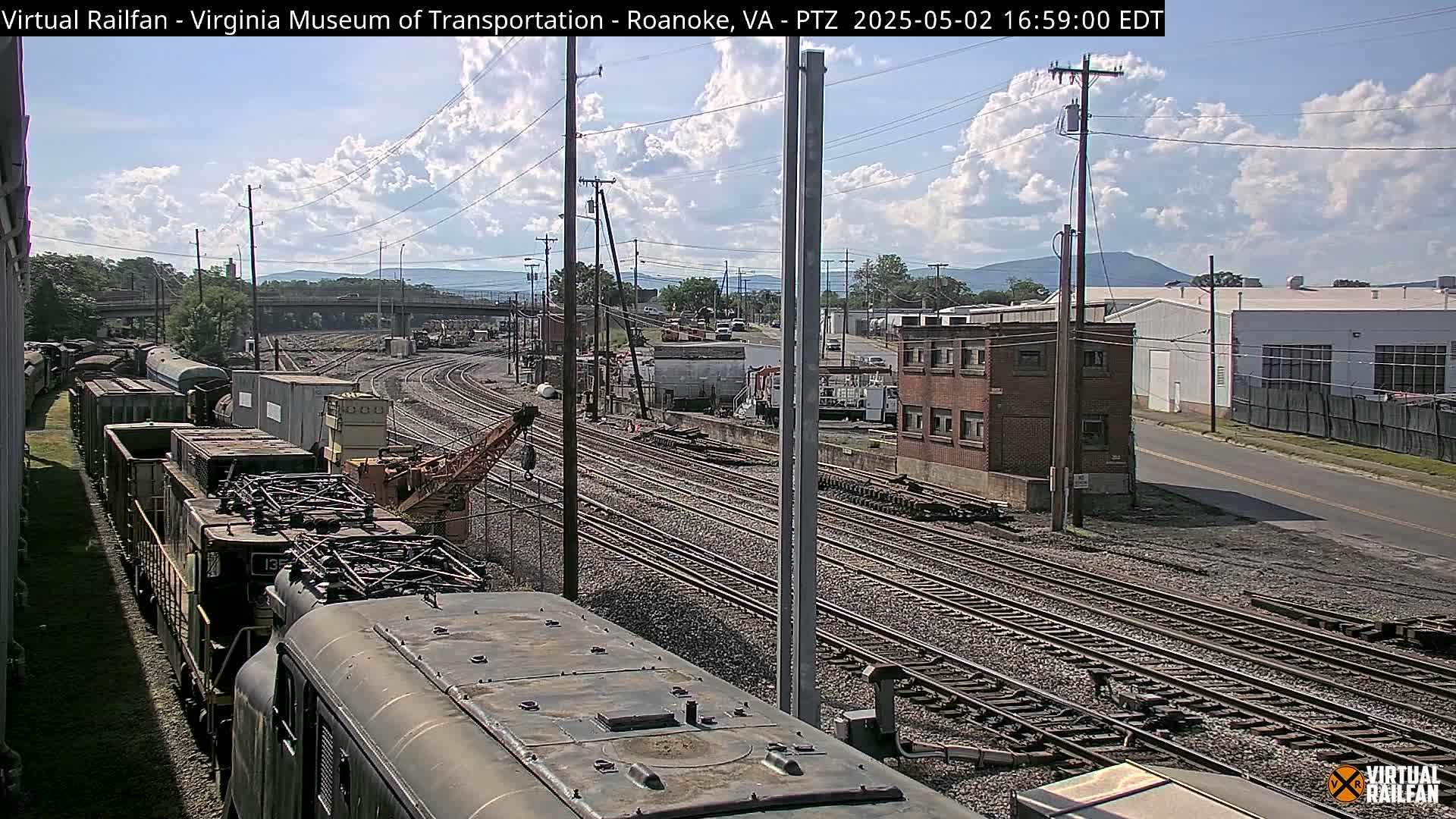 A partly cloudy day reveals a railroad yard with multiple train cars and tracks, a crane, and industrial buildings in the background.