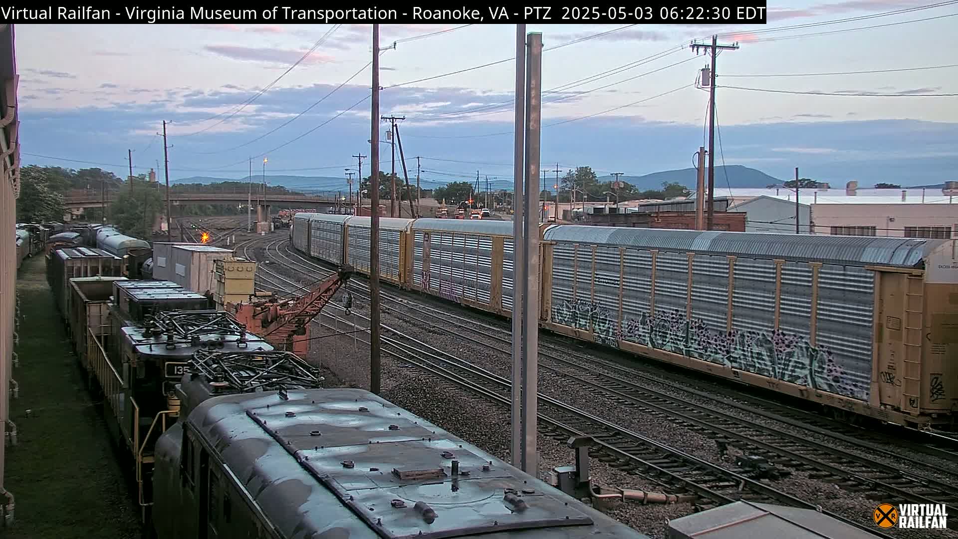 Two freight trains are visible on parallel tracks under a partly cloudy sky, with hills in the background.