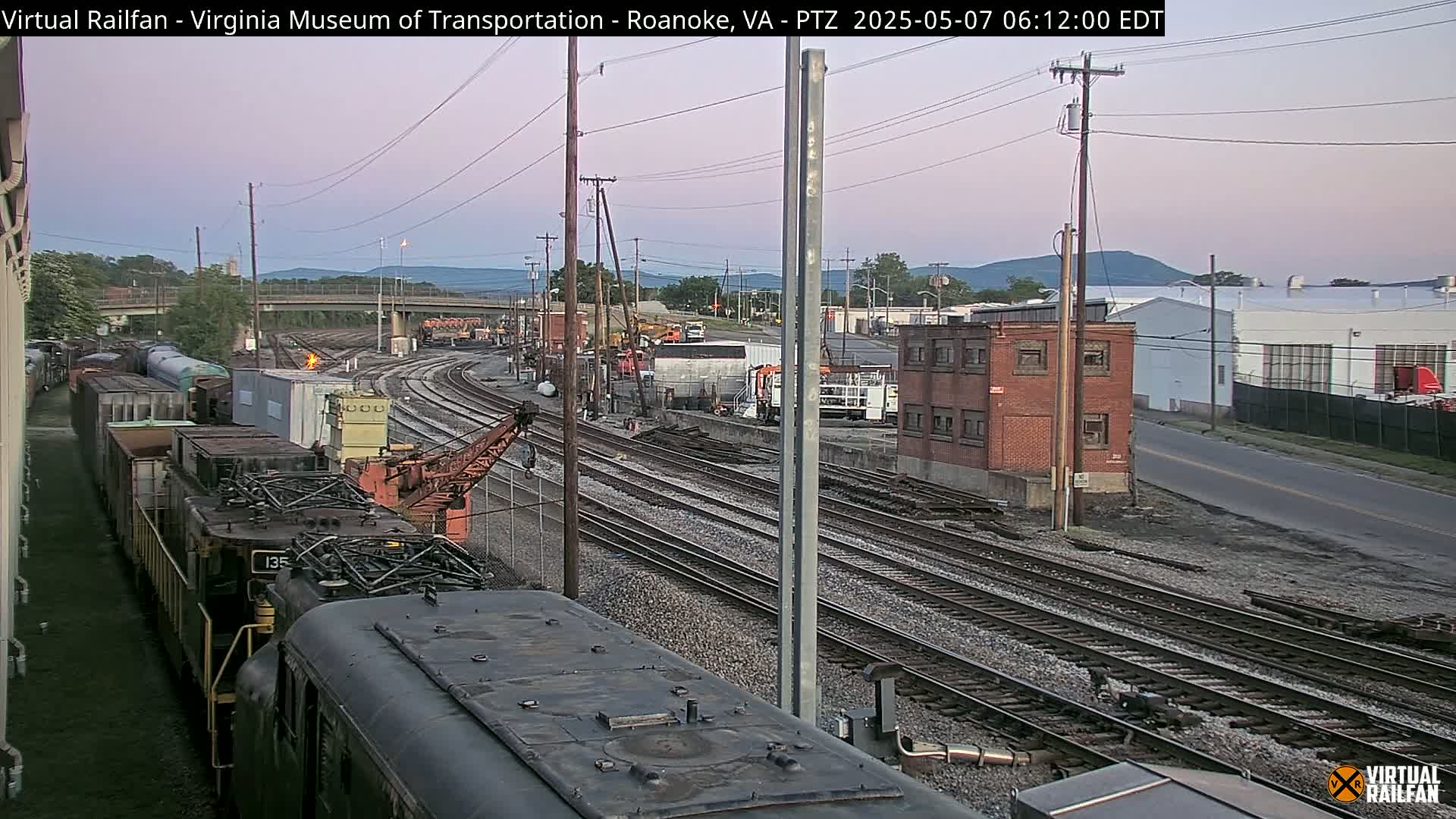 A twilight view of a railway yard with multiple train cars and tracks, a crane, and industrial buildings in the background.