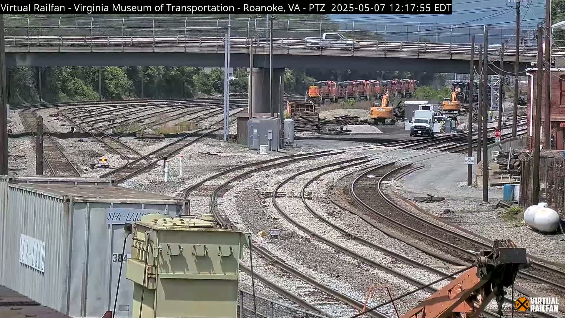 A sunny day reveals a complex railway yard with numerous train tracks curving under a bridge, several locomotives, and construction equipment.