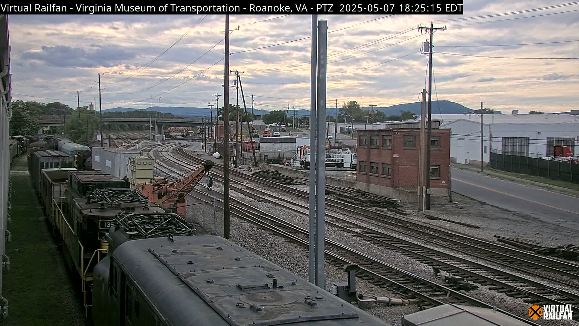 A partly cloudy evening scene shows multiple train tracks with several rail cars and locomotives, including a crane, near industrial buildings and a roadway, with mountains in the background.