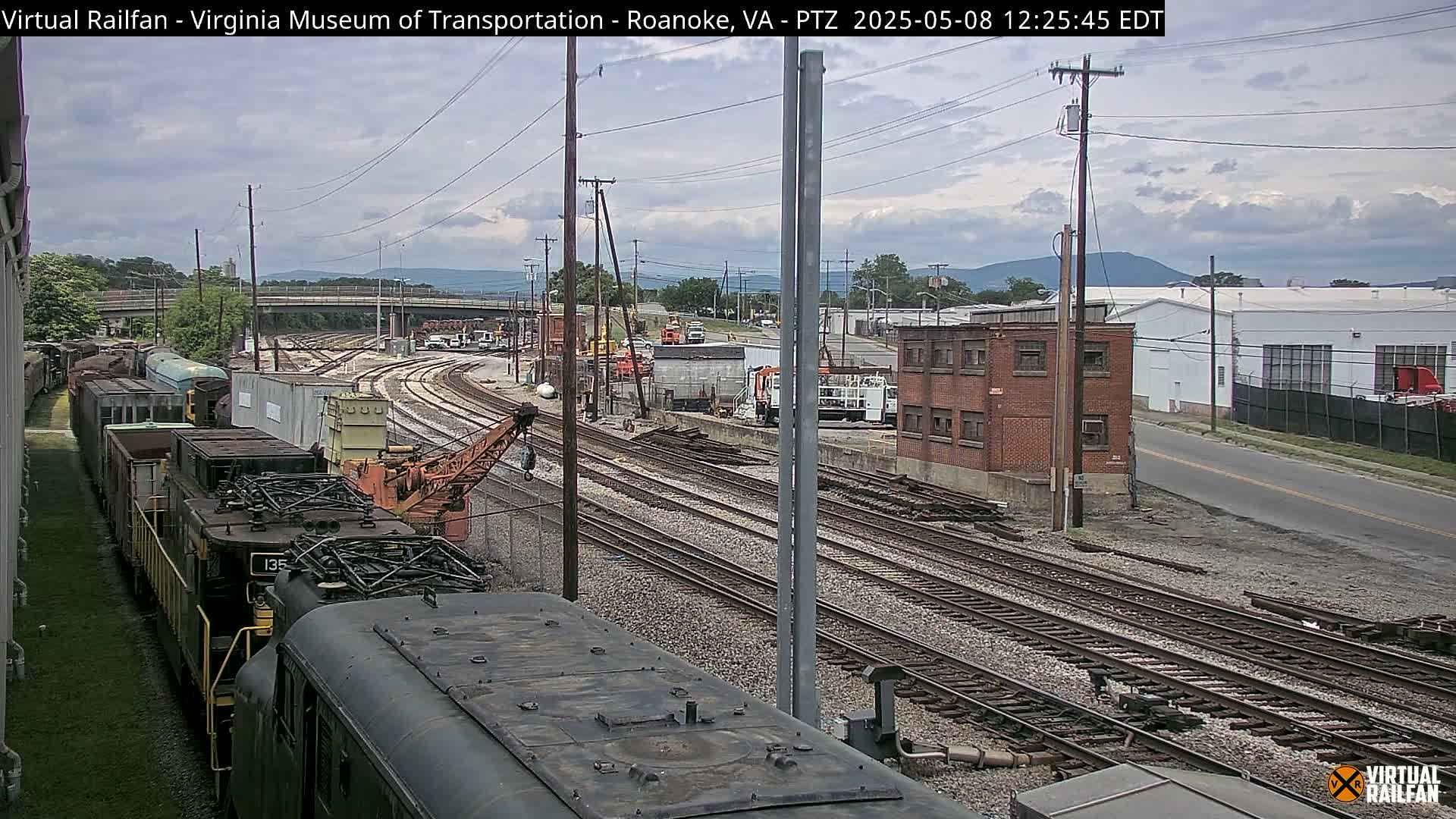 Under a partly cloudy sky, a train sits on a set of railway tracks next to a railway yard with several buildings and other trains.