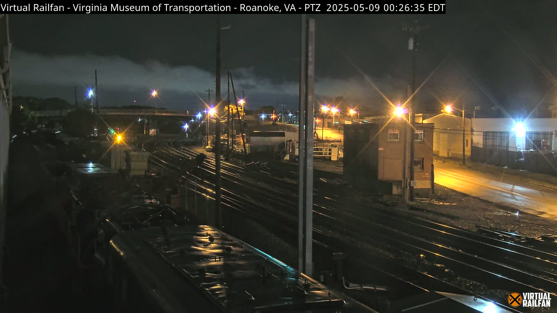 A nighttime view of a railroad yard with multiple tracks, illuminated by streetlights and the faint glow of buildings under a cloudy sky.